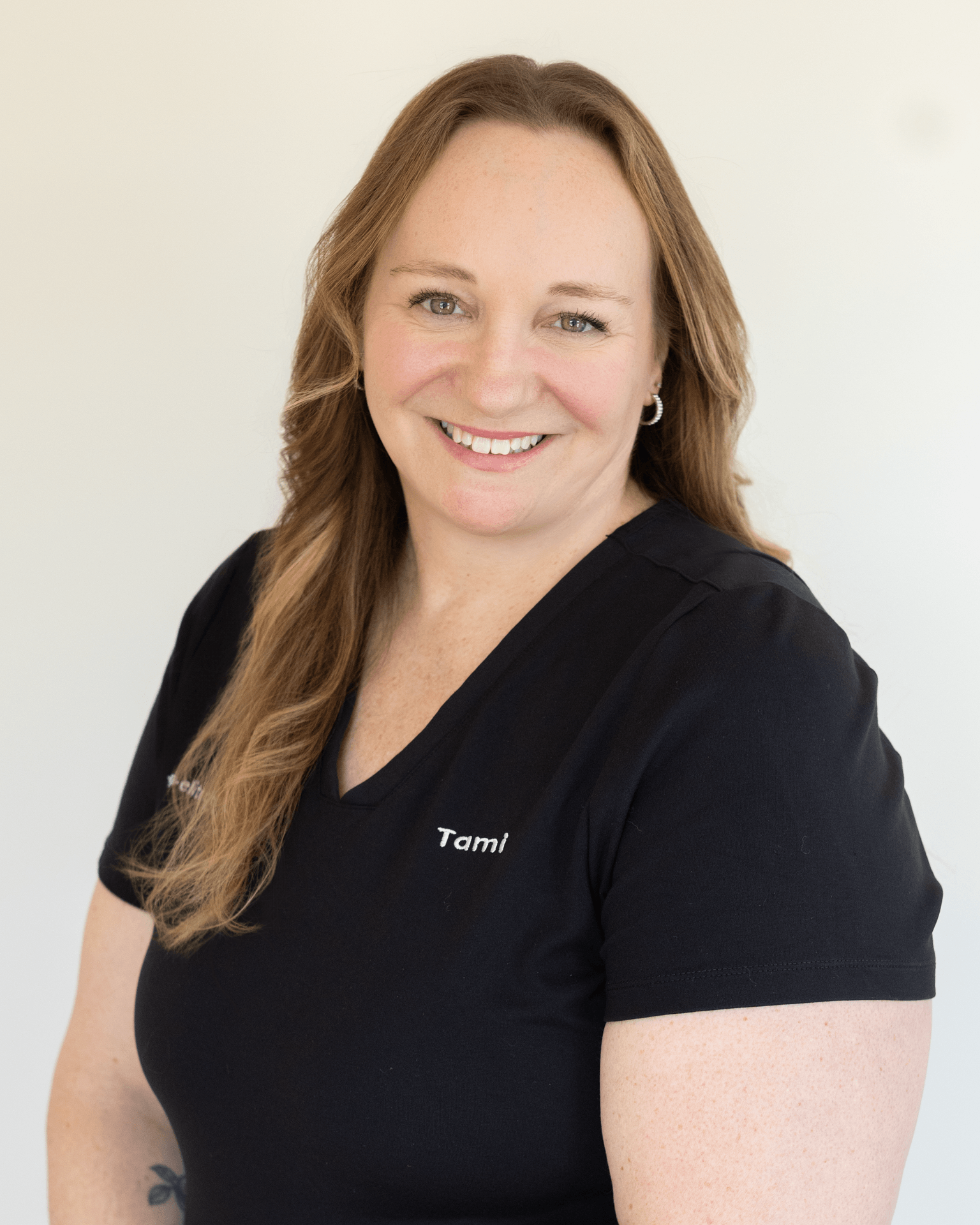 Close-up portrait of a smiling woman with long red hair, wearing a black shirt that says 'Tami', against a plain white background.