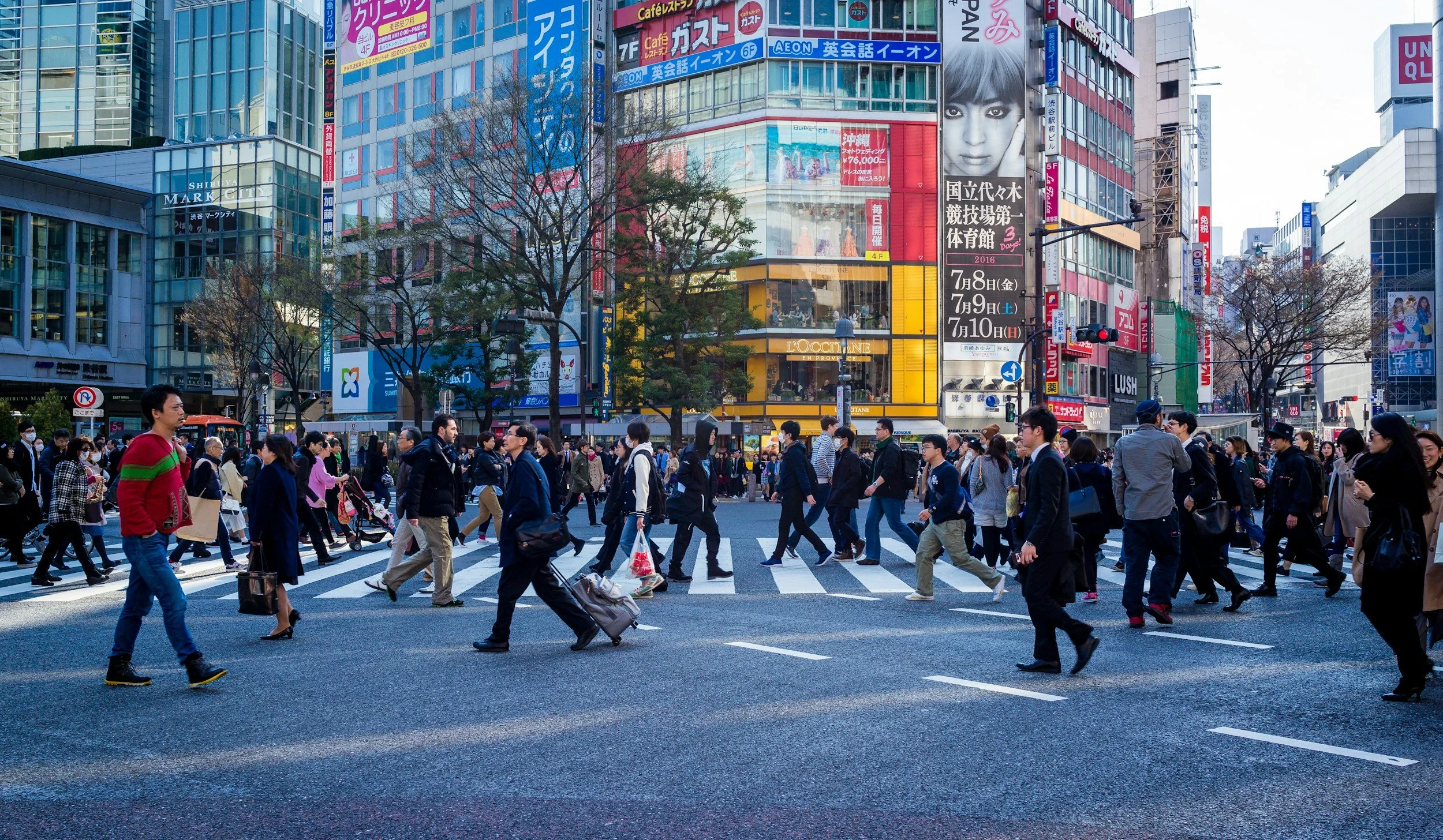 A busy intersection in an urban city, with many pedestrians crossing the street surrounded by tall, colorful buildings with billboards and advertisements.