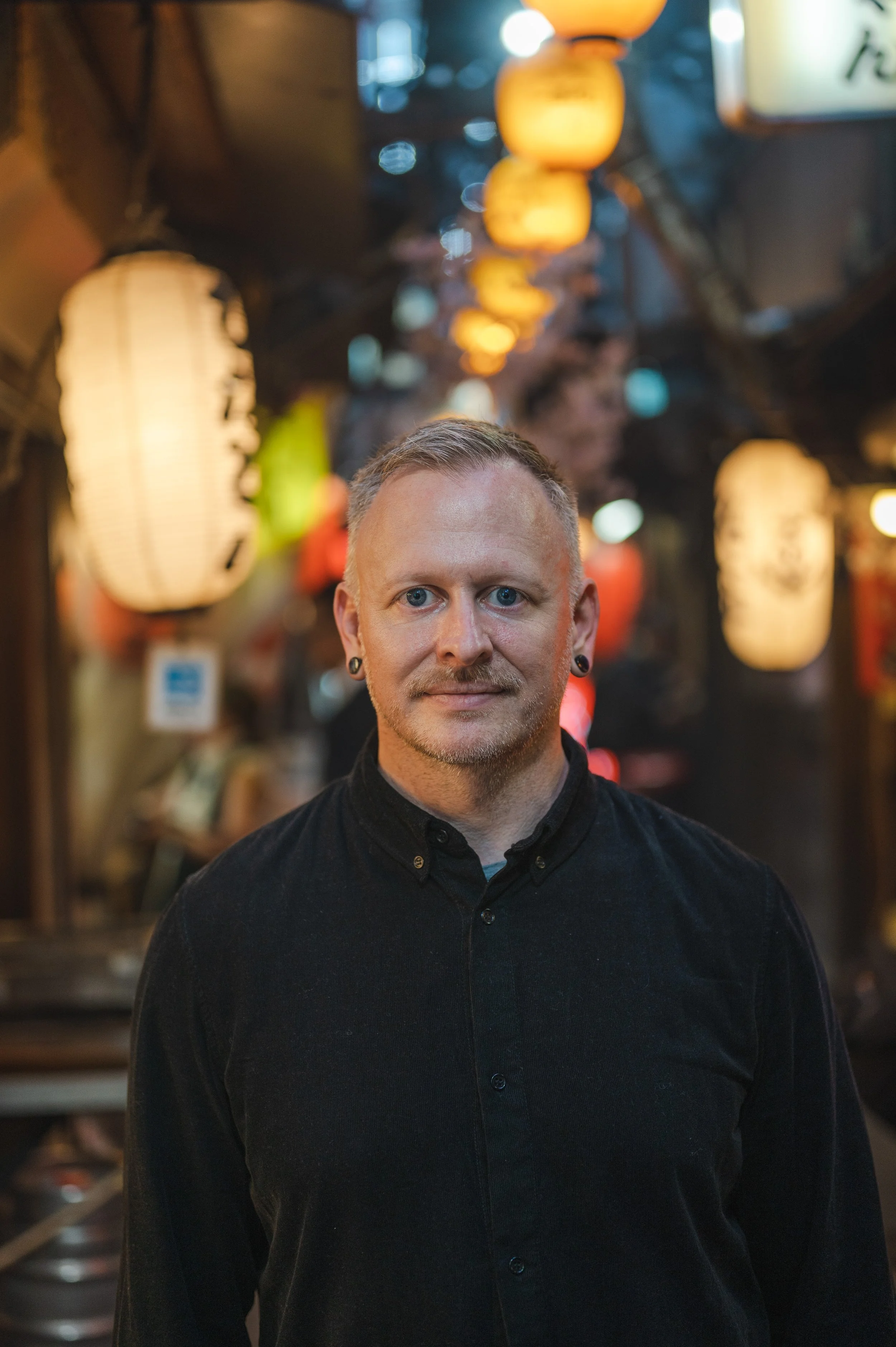 Image of Josh LaMar with short blond hair and earrings, wearing a black shirt, standing in a street in Tokyo decorated with hanging lanterns and colorful lights at night.