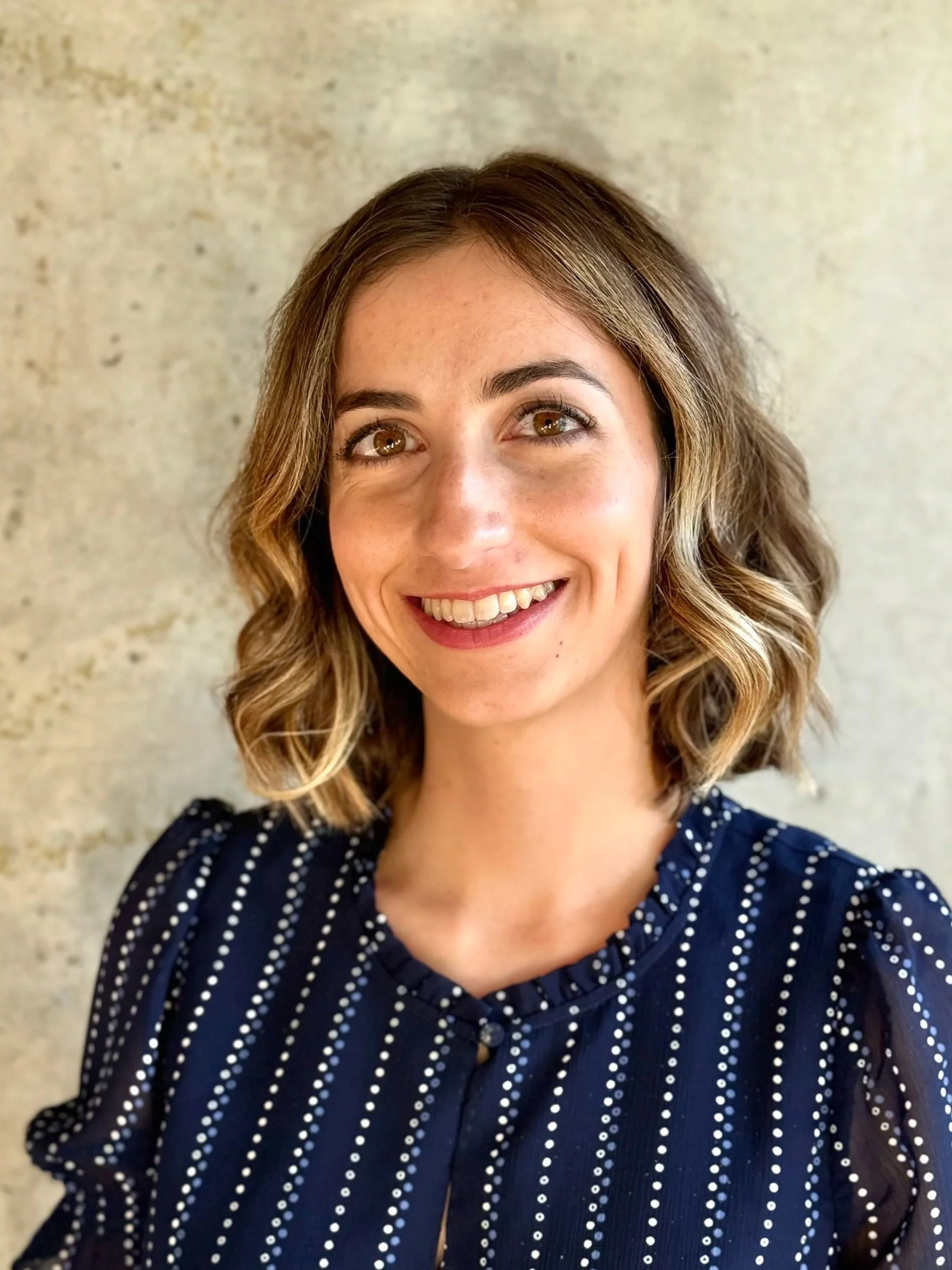 A young woman with short wavy hair smiling, wearing a navy blue blouse with white polka dots, standing against a textured beige wall.