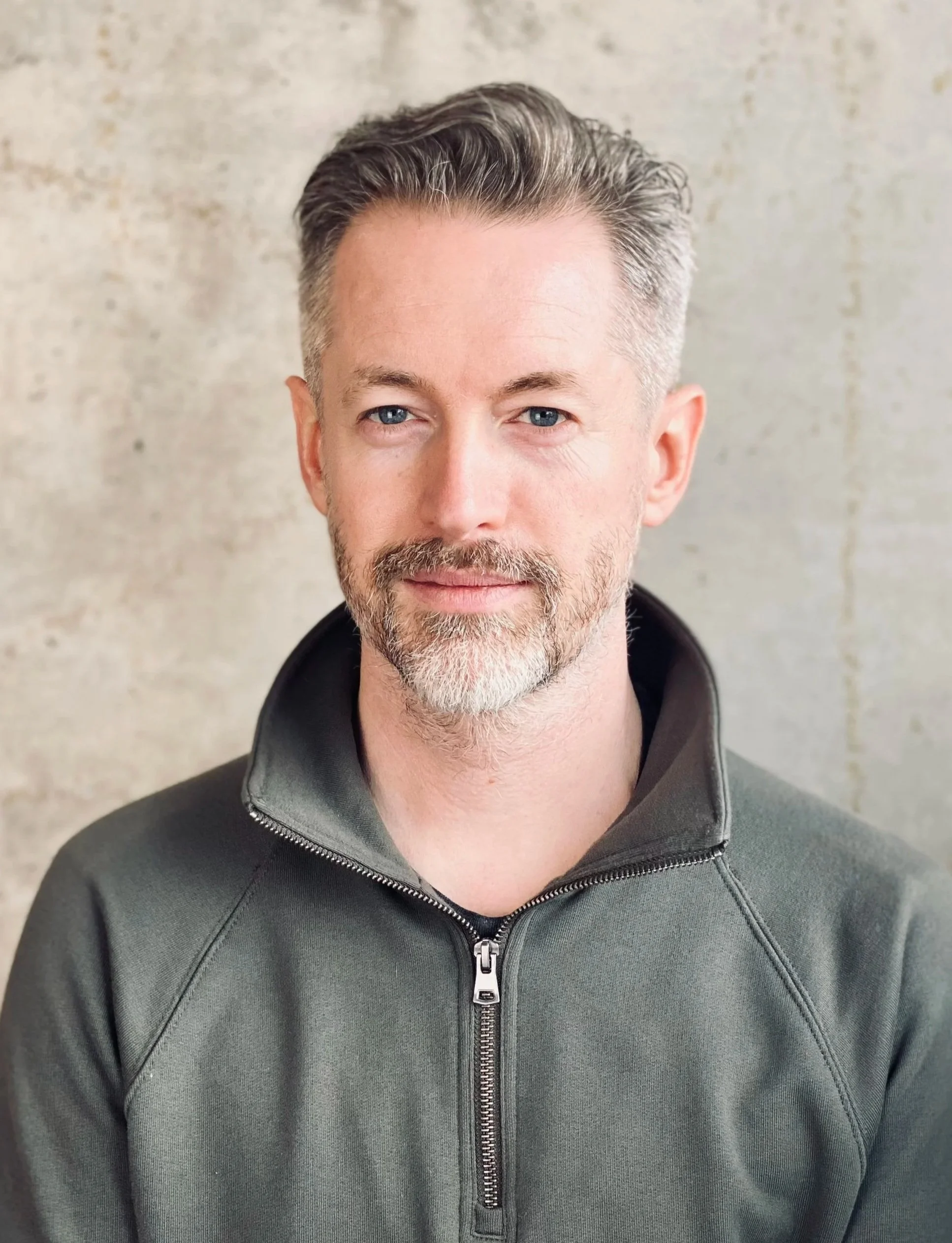 A middle-aged man with short, graying hair and a beard, wearing a dark green zip-up sweater, stands in front of a textured, light-colored wall.