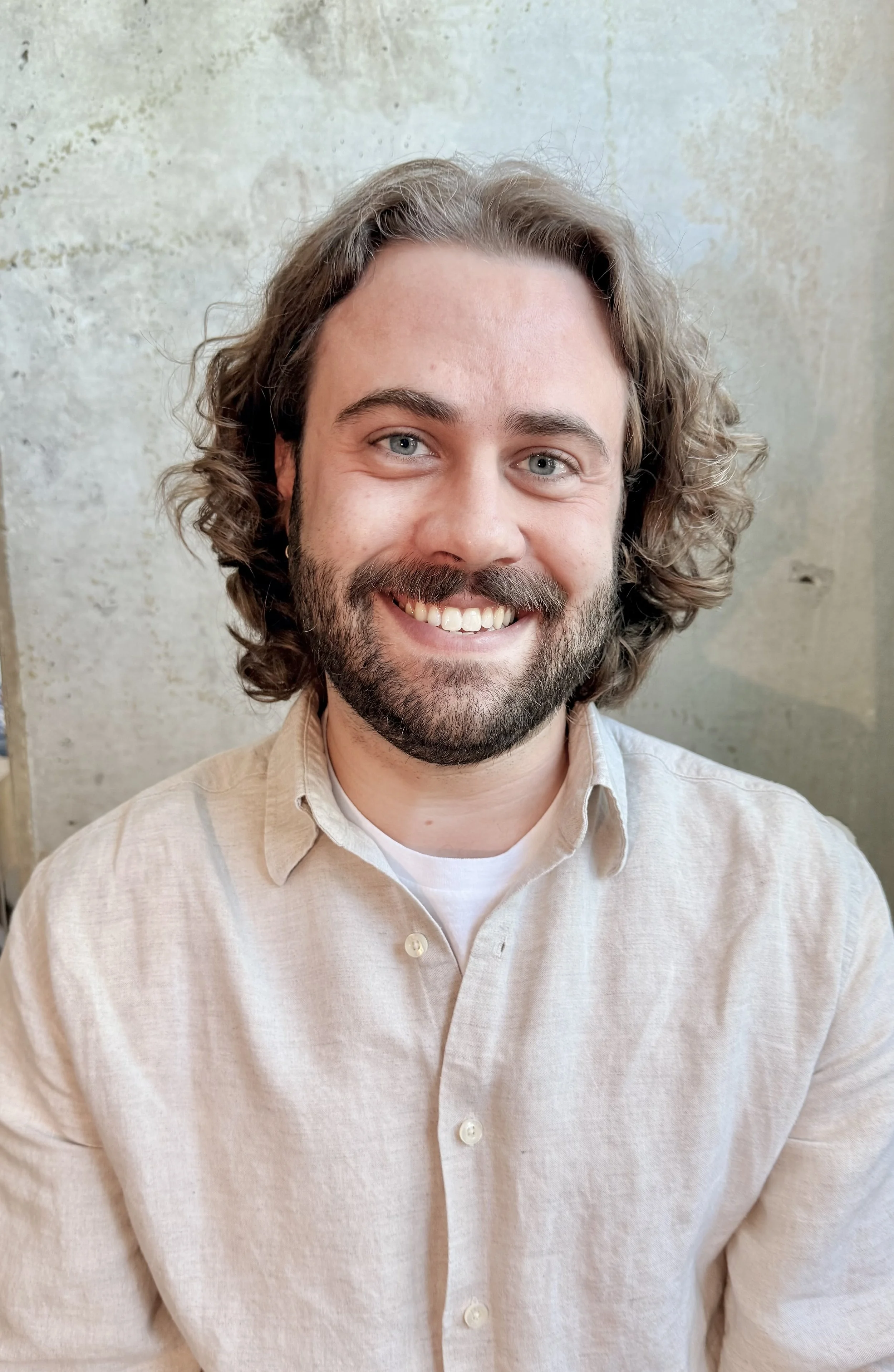 A smiling man with curly brown hair, a beard, and blue eyes, wearing a striped button-up shirt, standing against a textured beige wall.