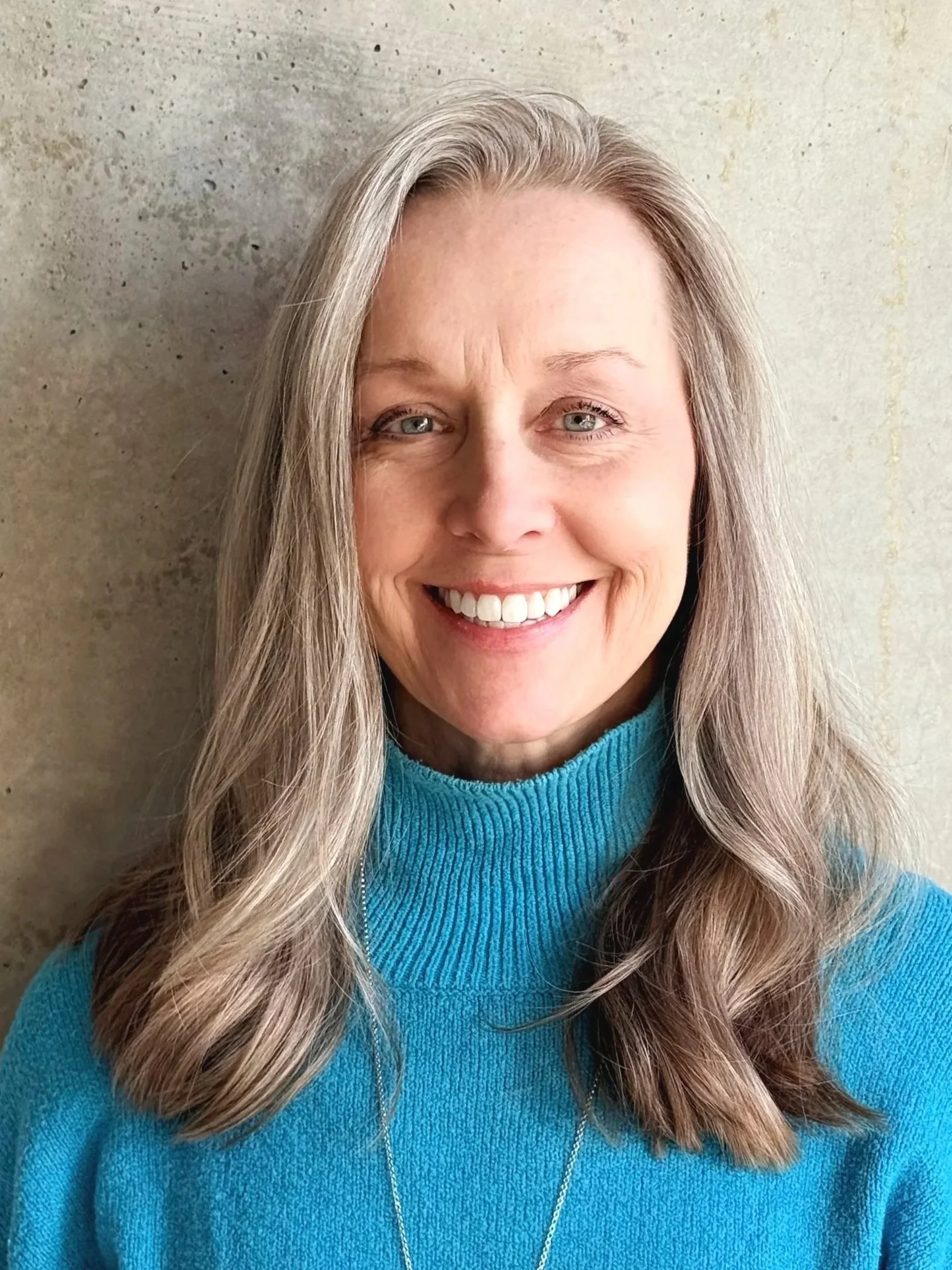 A middle-aged woman with long gray hair and blue eyes, smiling, wearing a bright blue turtleneck sweater and a silver necklace, standing against a plain, textured beige wall.