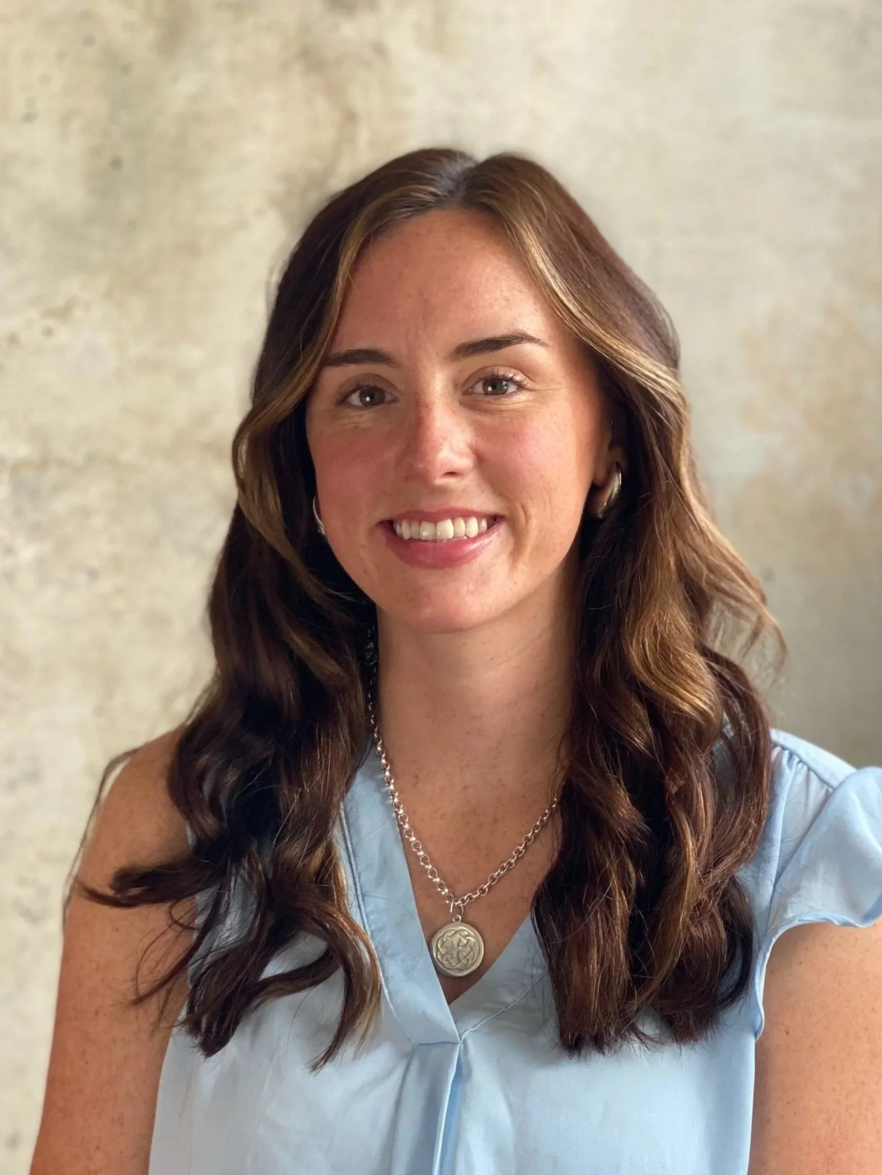 A woman with long, wavy brown hair wearing a light blue blouse and a silver necklace with a round pendant, smiling at the camera in front of a neutral background.
