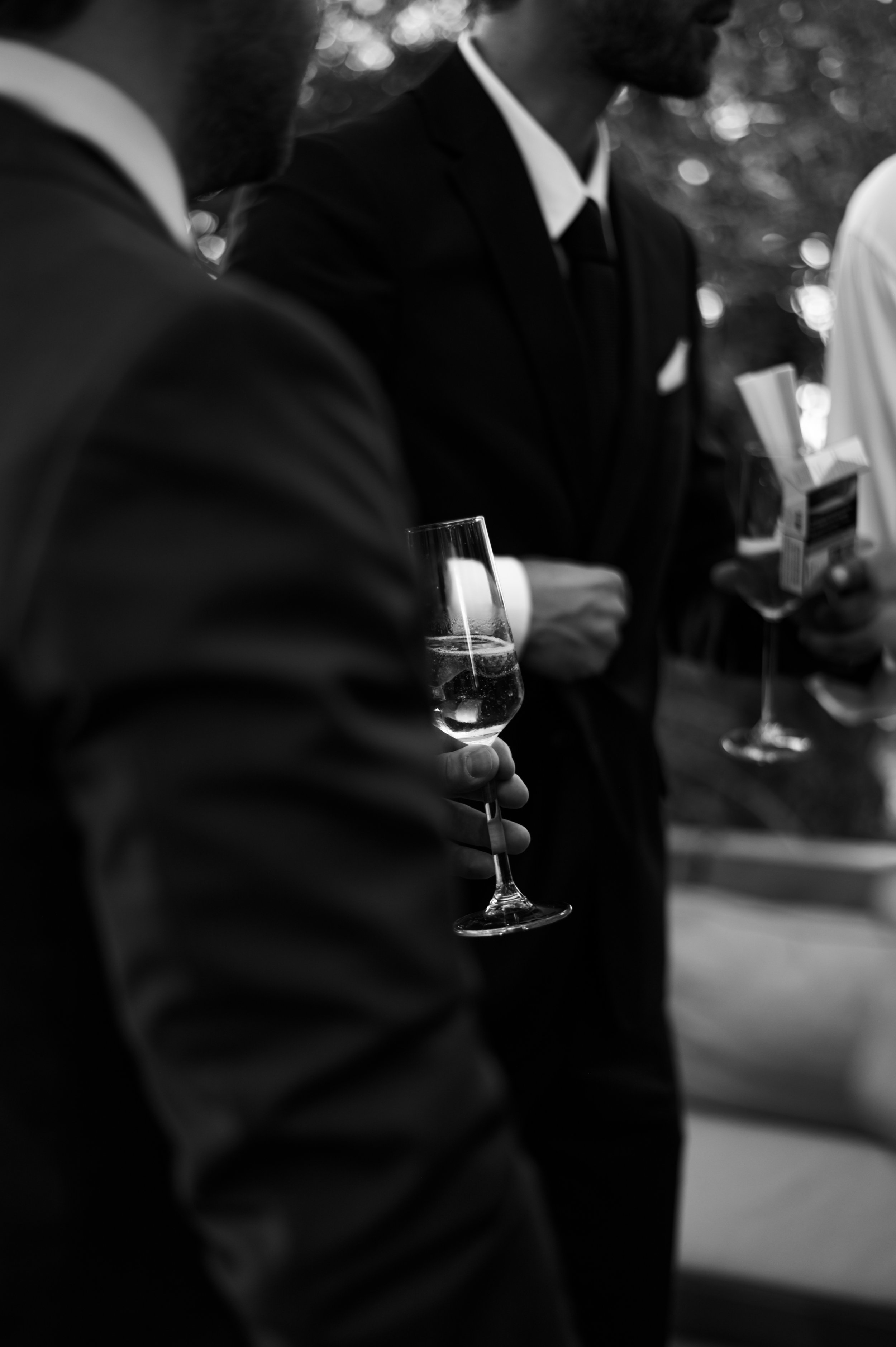 Men in formal attire holding drinks and a pack of cigarettes at a social gathering, black and white photo.