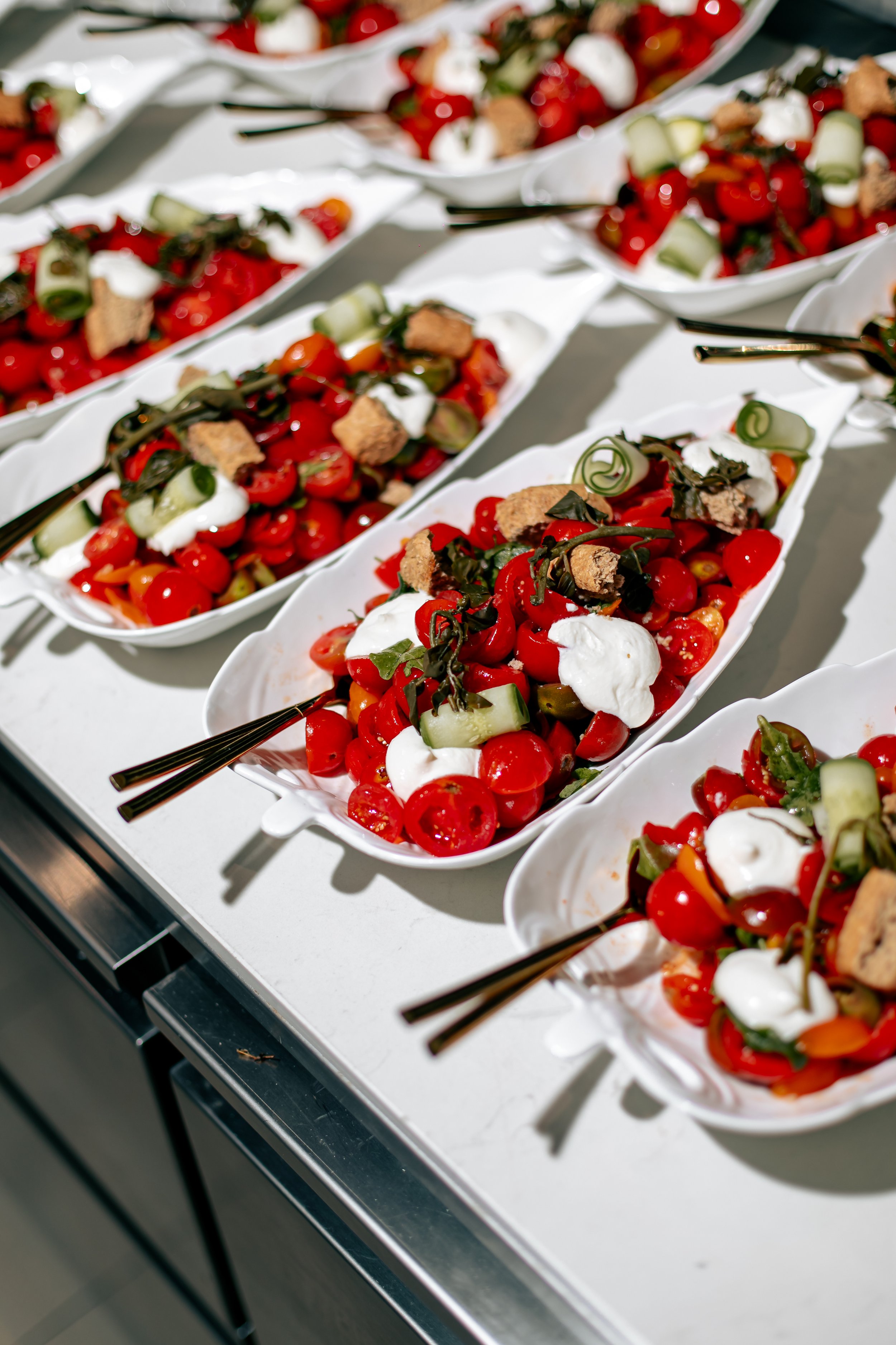 A display of several white oval bowls filled with cherry tomato salads, topped with small pieces of bread, dollops of white cheese, and curled cucumber slices, arranged on a white table.