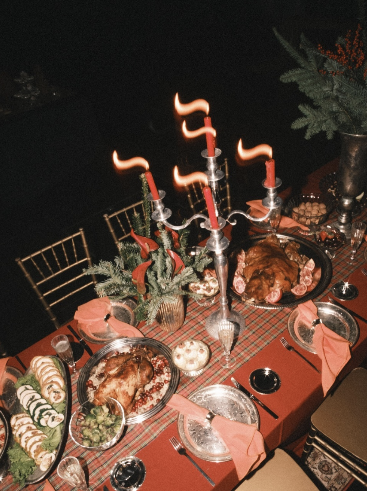Festive dinner table with roasted turkey, side dishes, holiday decorations, a candelabra with lit candles, and a plaid tablecloth.