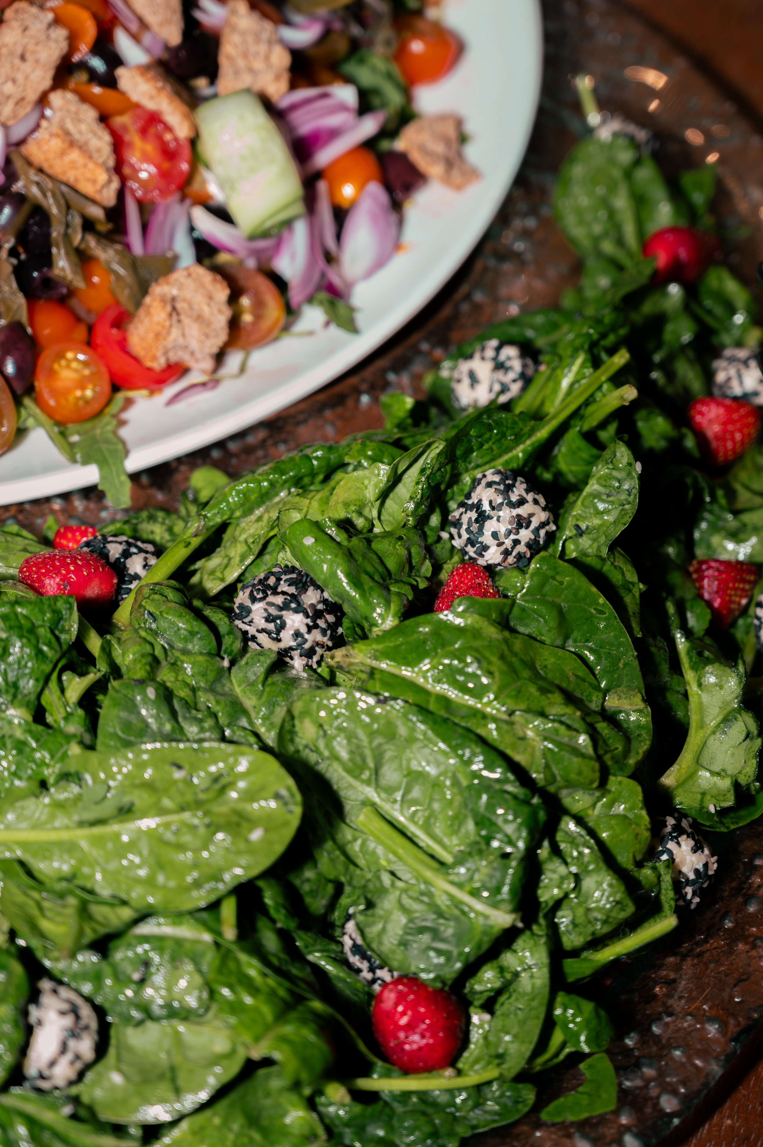 Close-up of a fresh spinach salad with strawberries and black and white sesame seed balls, alongside a separate bowl of vegetable salad containing cherry tomatoes, cucumbers, red onion, and croutons.