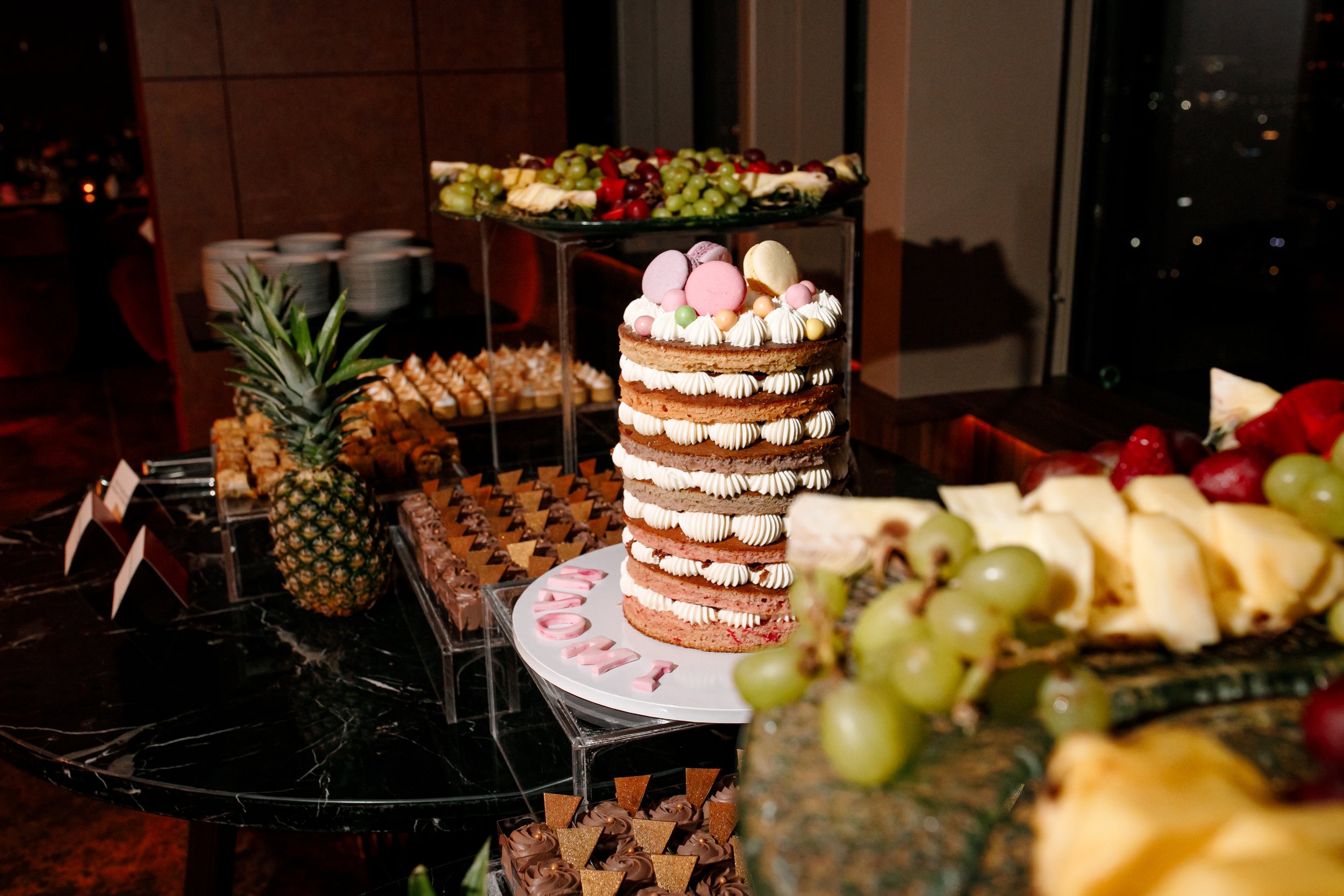Sweet table with a pineapple, layered cake with pink, white, and green macarons, grapes, pineapple, and assorted pastries, in a nighttime cityscape setting.