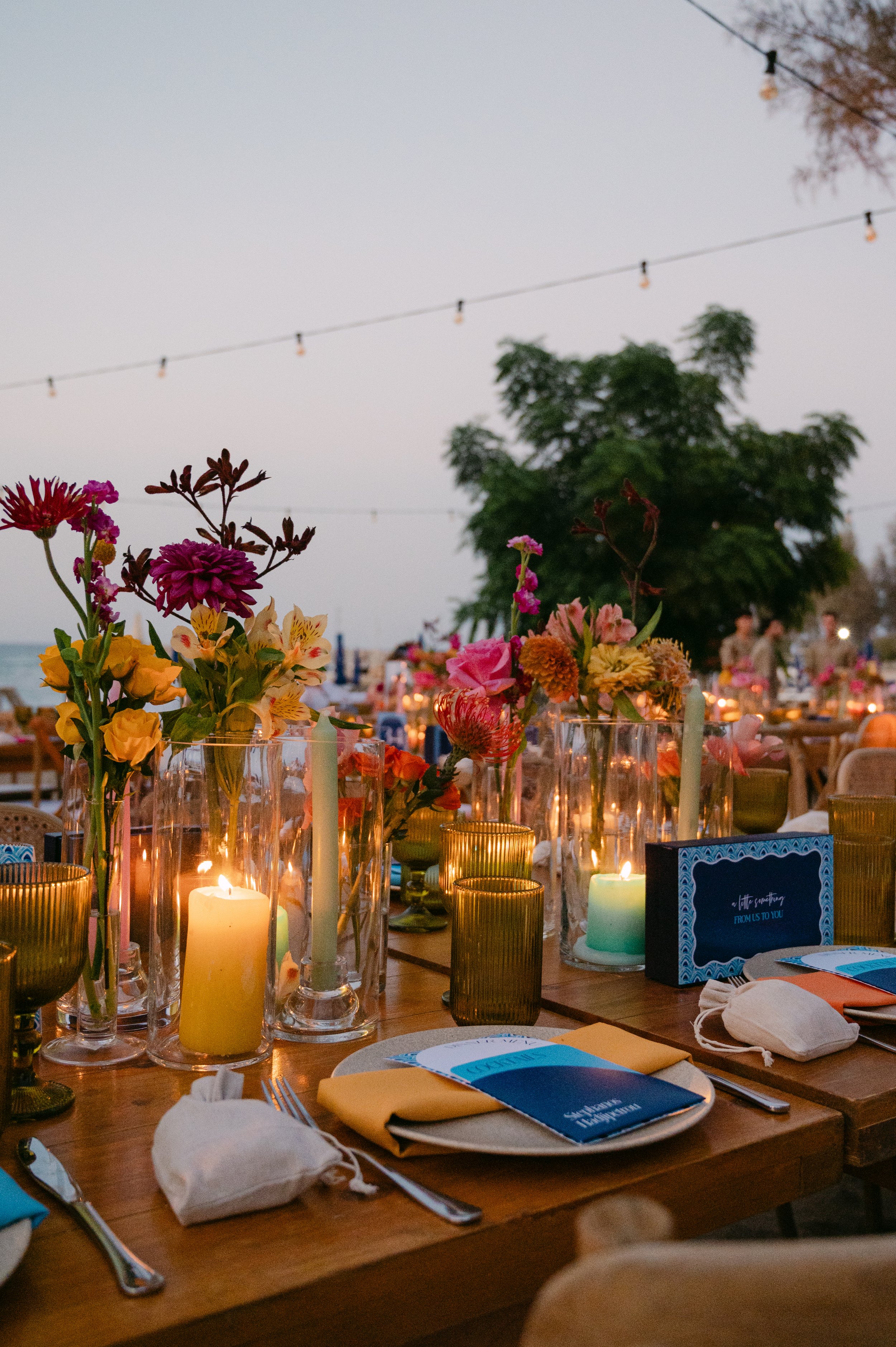 Nighttime outdoor dining table decorated with colorful flowers and lit candles, with string lights overhead and trees in the background.