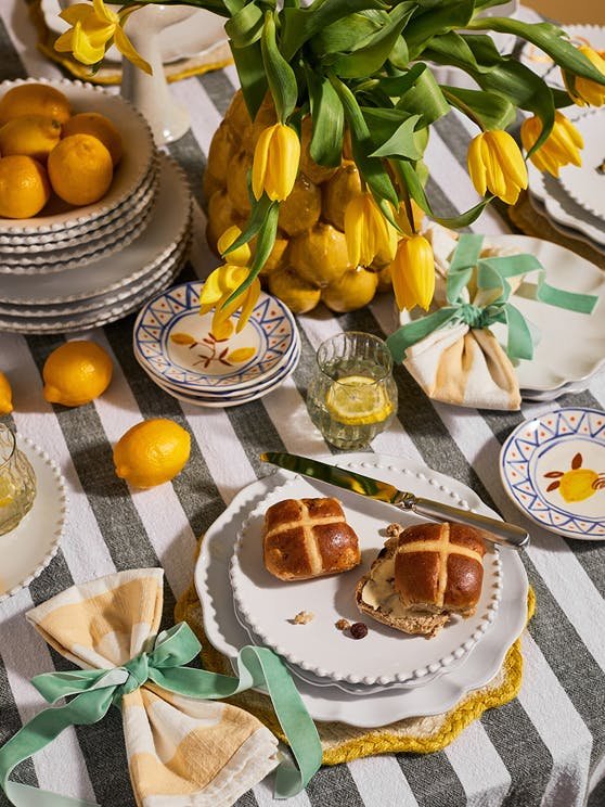 Easter table setting with yellow tulips in a vase, lemons in a bowl, and a plate with hot cross buns. Decorated napkins, colorful plates, and a glass of lemonade are also on the table with a striped tablecloth.