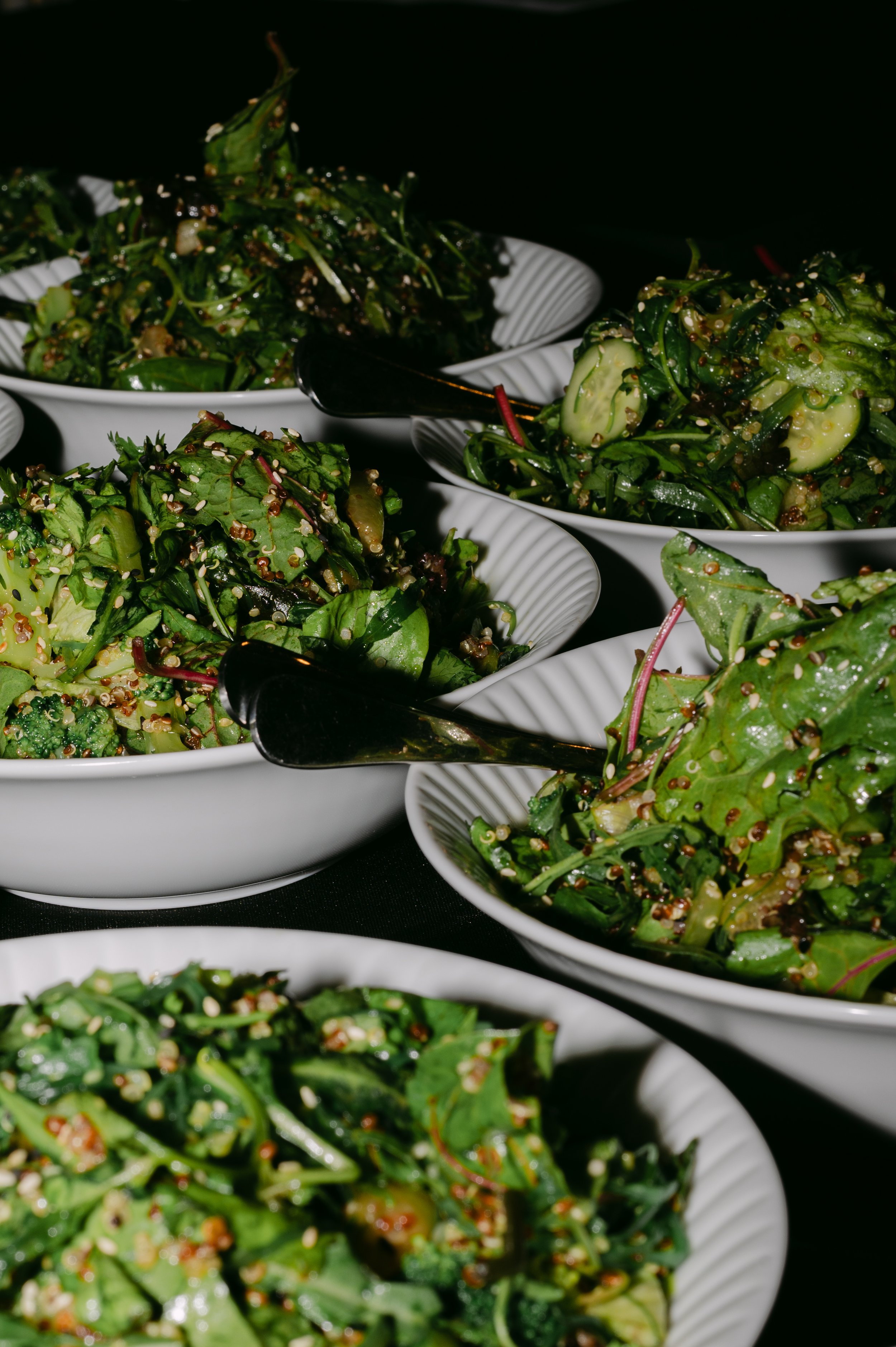 Multiple bowls of mixed green salad with quinoa, cucumbers, and greens, served with black utensils on a black background.