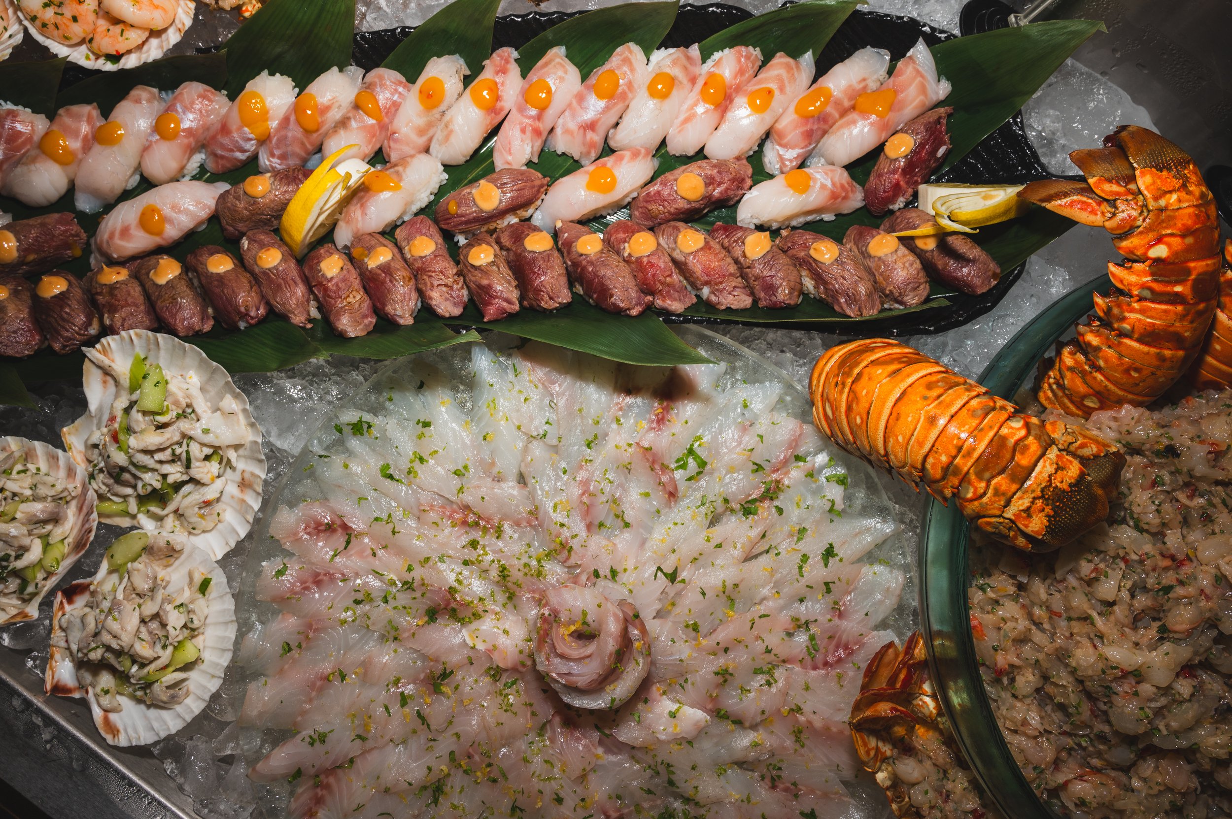 A seafood platter featuring various types of raw fish, shrimp, lobster tails, and sashimi arranged on a bed of ice, garnished with lemon and herbs.