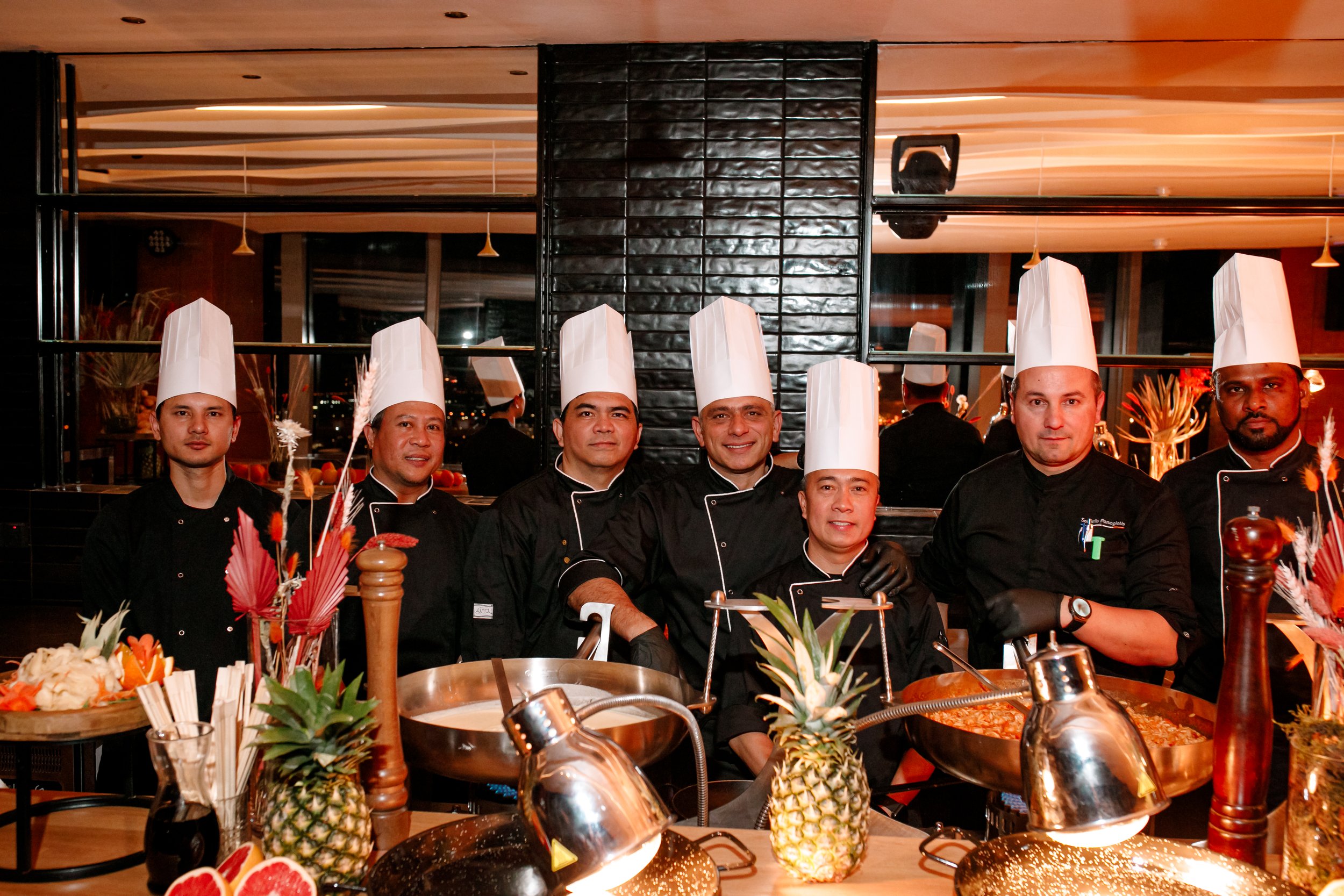 Group of professional chefs in black uniforms and white chef hats posing in a restaurant kitchen with tropical fruits and cooking equipment in the foreground.