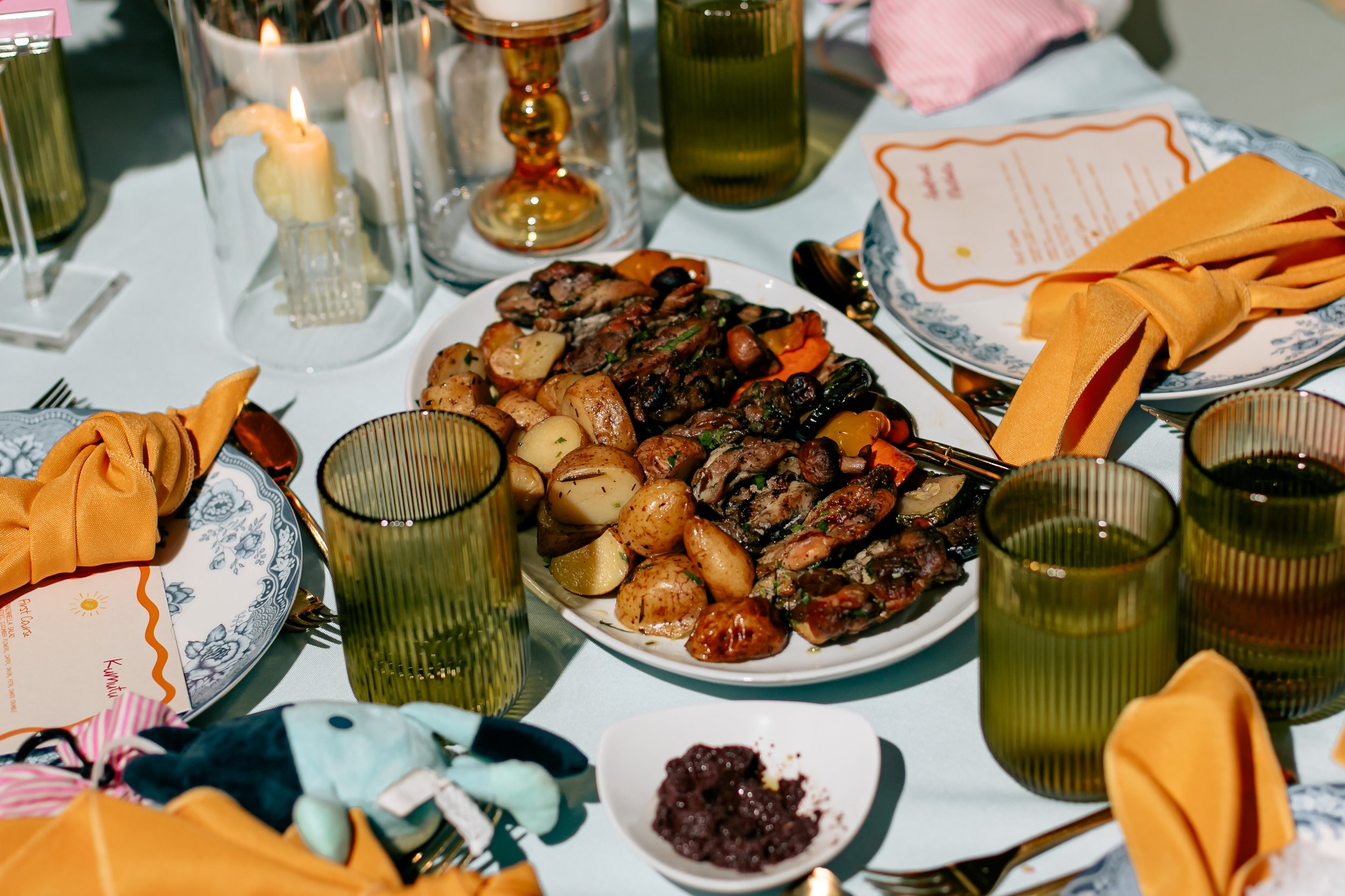 A dinner table set with a platter of roasted vegetables and meats, surrounded by green tinted glasses, yellow napkins, and place settings with menu cards.