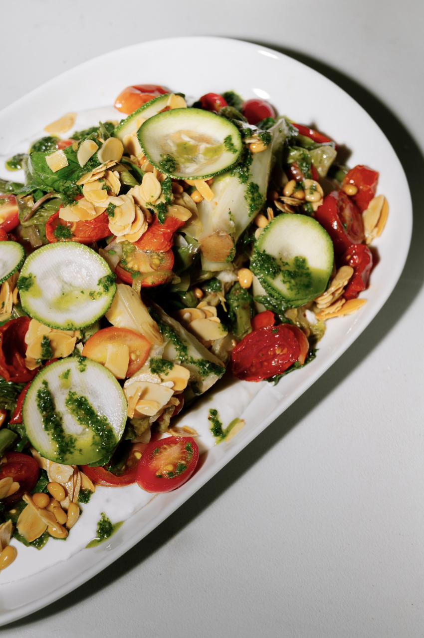 A fresh vegetable salad with sliced cucumbers, cherry tomatoes, chopped greens, toasted almond slices, and a green herb dressing on a white oval plate.
