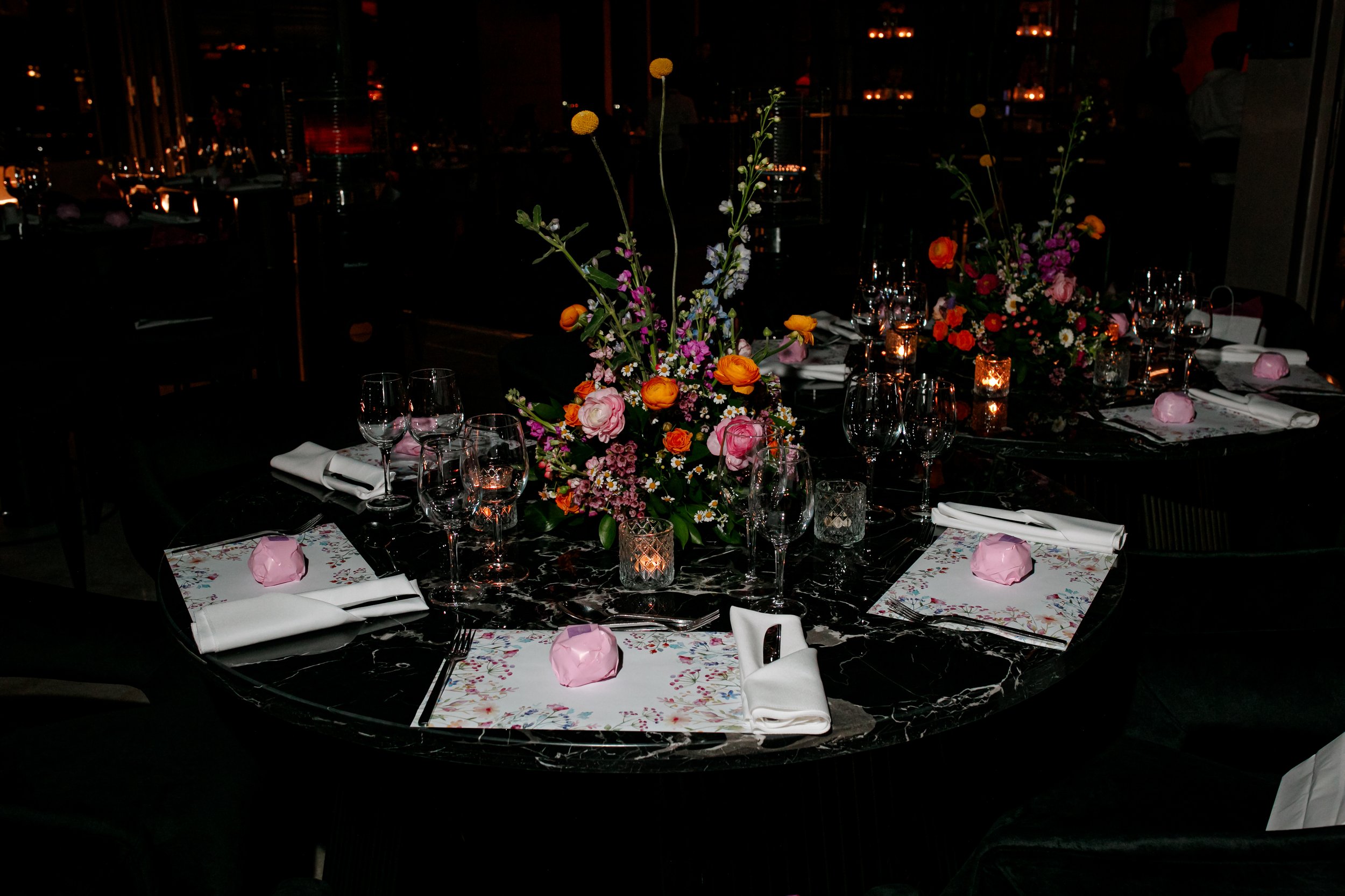 Elegant round table set for a formal dinner with floral centerpieces, wine glasses, candles, and napkins with pink paper-wrapped favors on a black marble table.