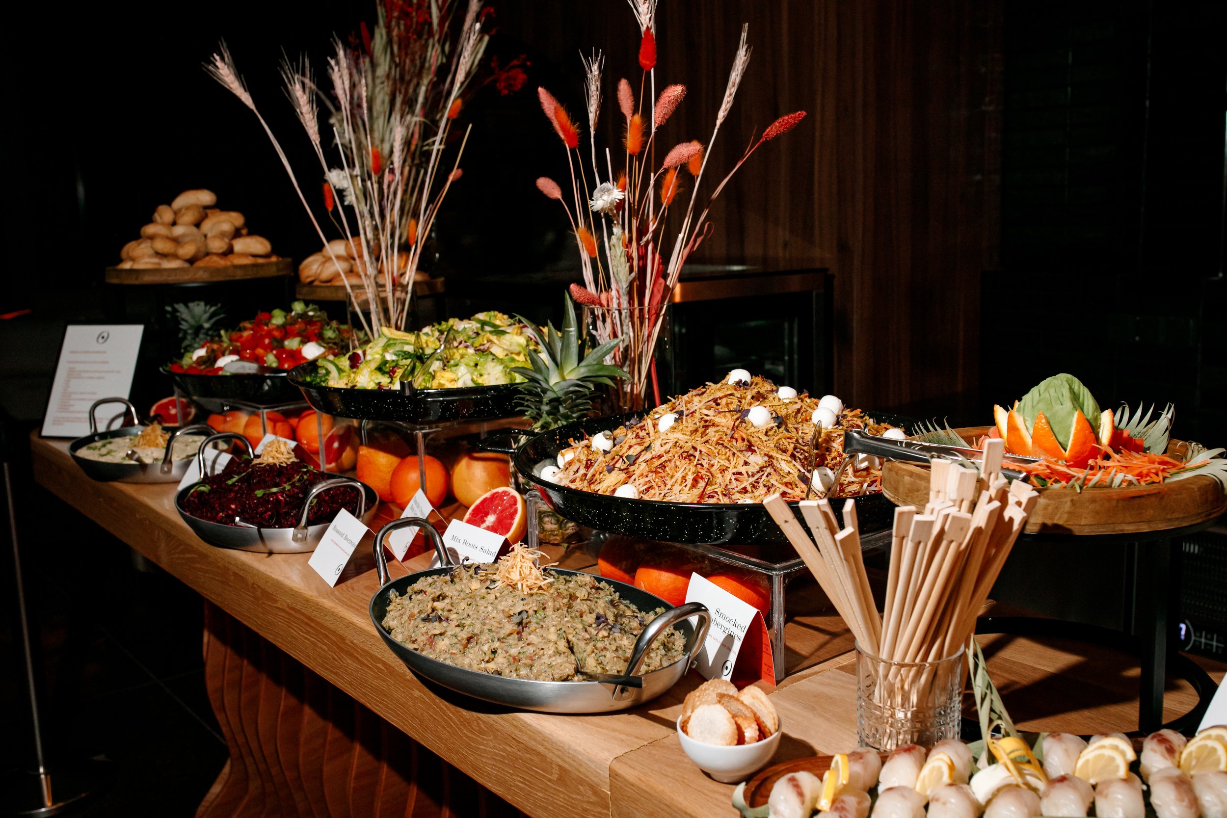 Buffet table with various salads and dishes, including mixed greens, shredded meat with eggs, beet salad, grain salad, and a decorative centerpiece with dried flowers and a pineapple, in a dimly lit room.