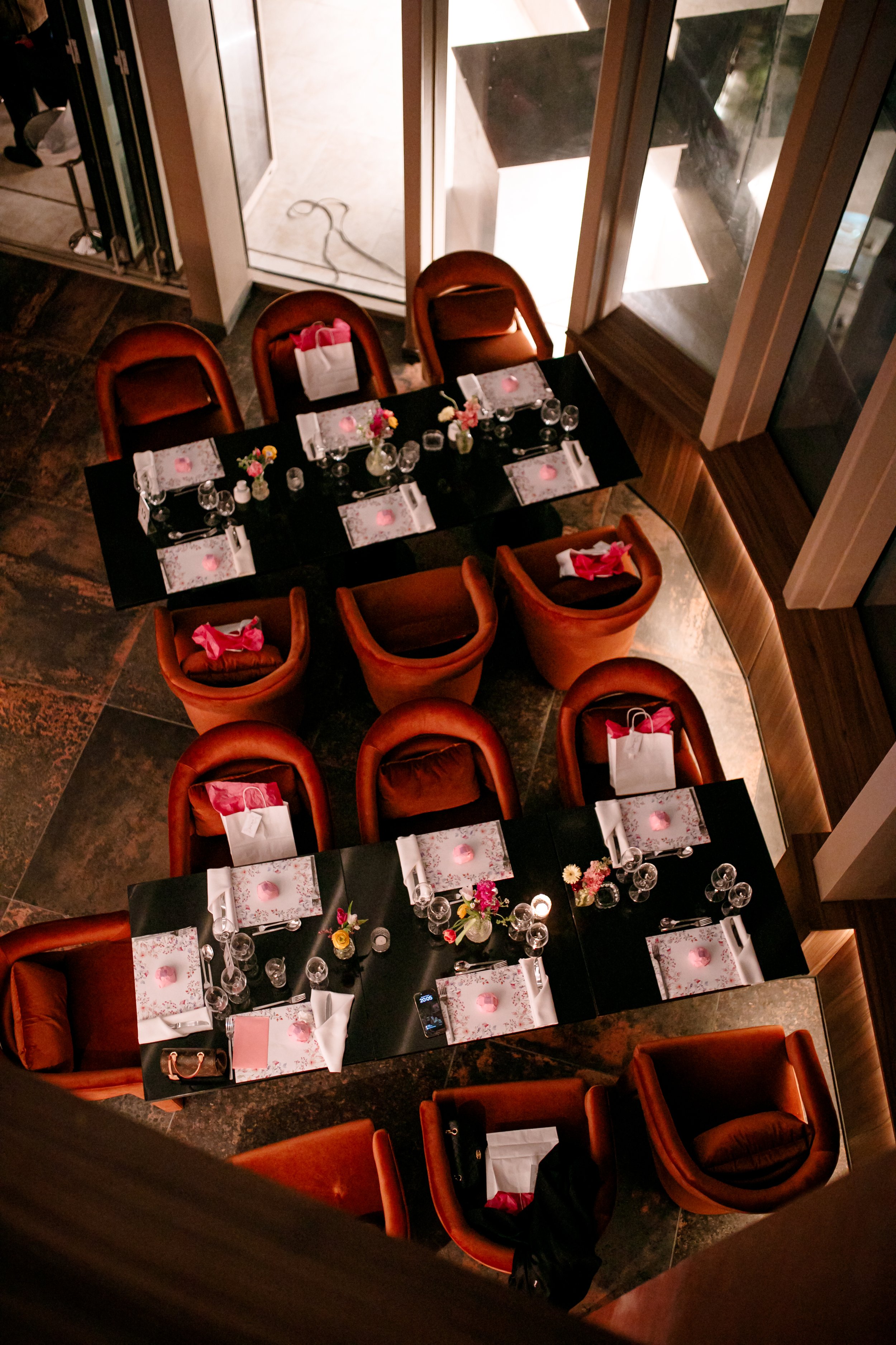 Top-down view of a dining table set for eight with floral centerpieces, candles, and place settings, surrounded by orange upholstered chairs in a warmly lit restaurant.