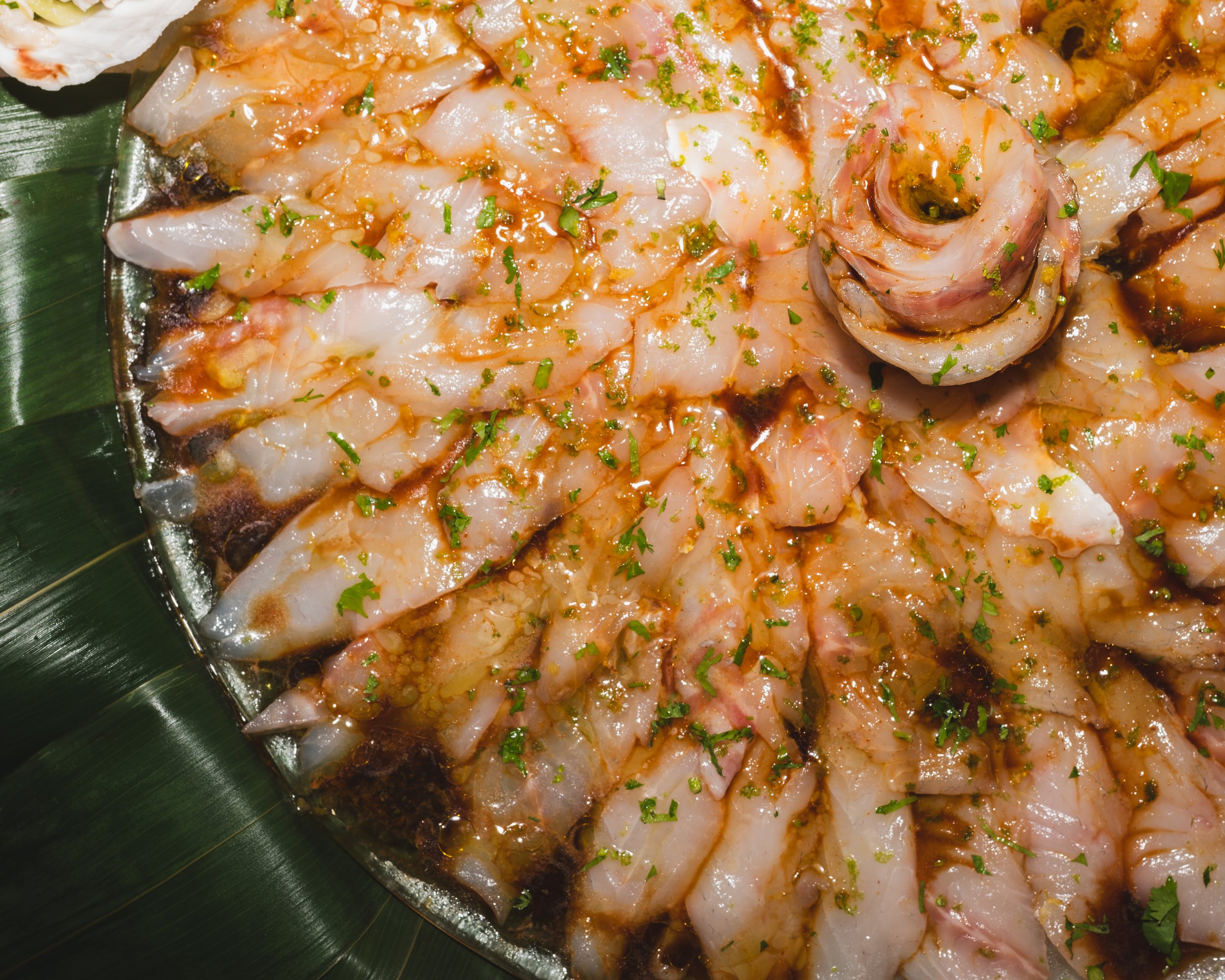 Raw fish fillets garnished with chopped herbs and drizzled with sauce on a round silver platter with green leaves around the edge.