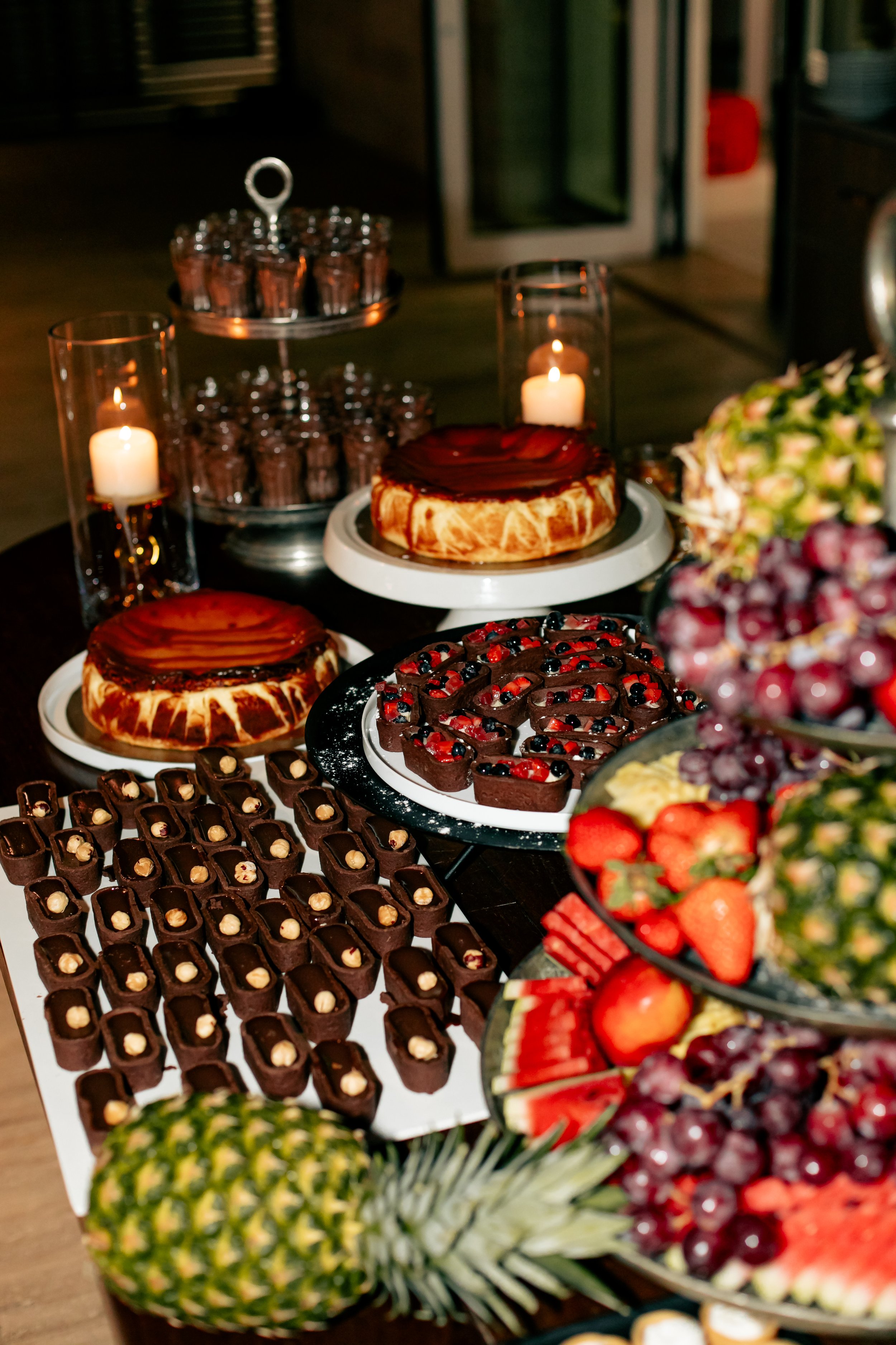 A variety of desserts and fruits on a table, including chocolates, cakes, strawberries, grapes, pineapples, and berries, with candles in the background.