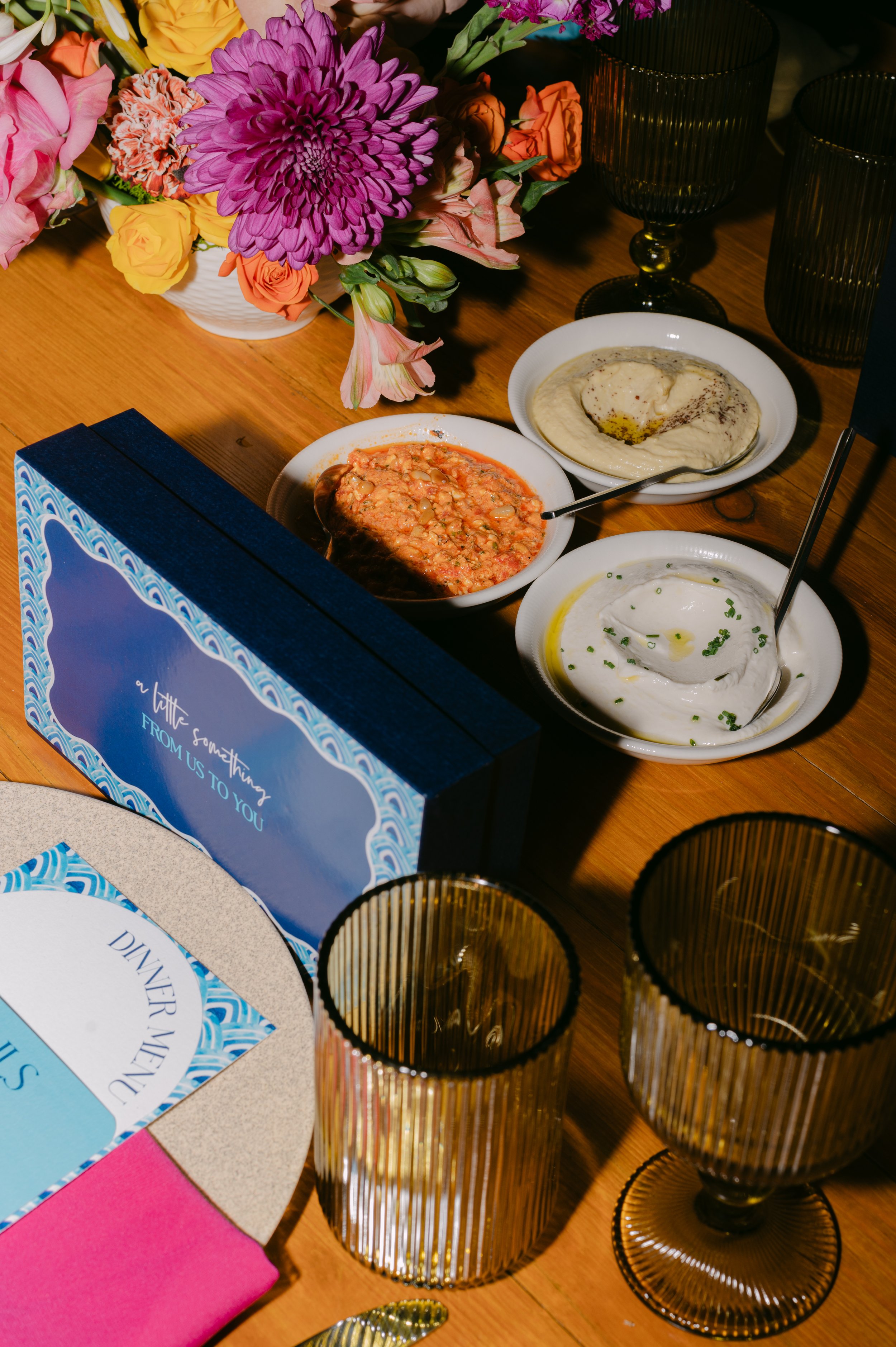 A wooden table with three bowls of creamy dips, a bouquet of pink, purple, yellow, and orange flowers, decorative amber glasses, a blue and white card with the message 'a little something FROM US TO YOU,' part of a pink napkin, and a menu labeled 'DI