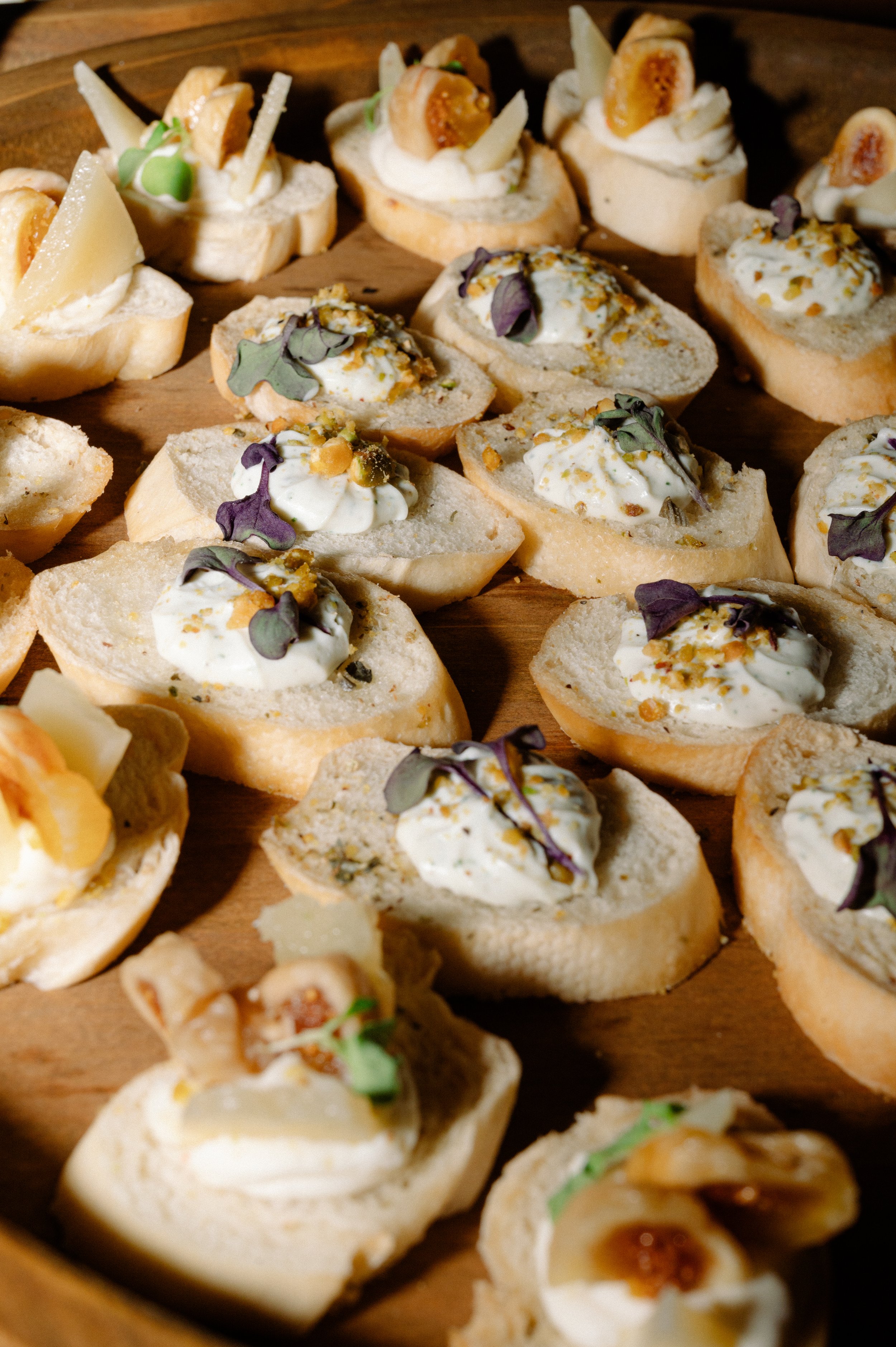 Assorted canapé appetizers on a wooden platter, topped with cheeses, microgreens, and chopped nuts.