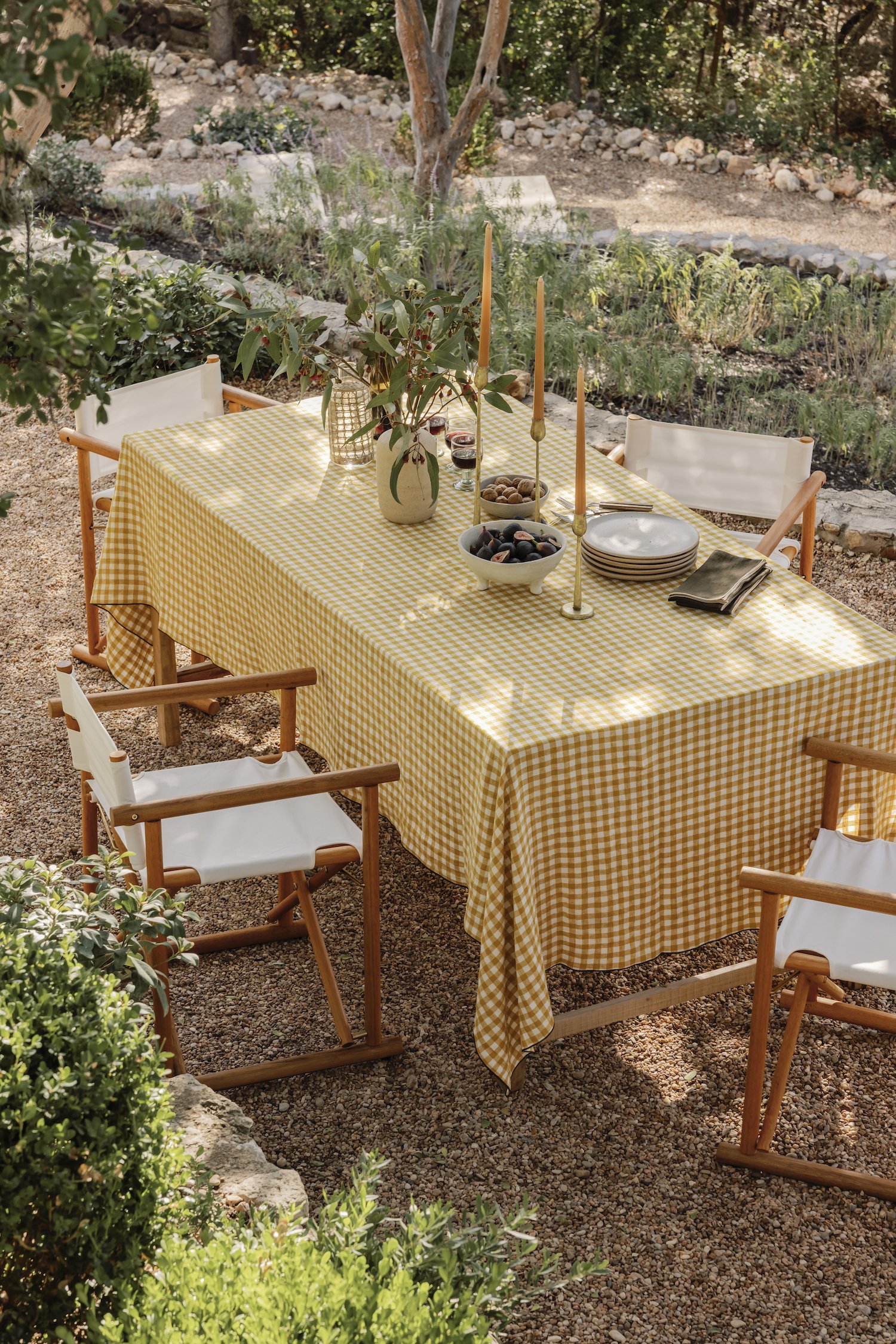 Outdoor dining table with a yellow checkered tablecloth, surrounded by wooden chairs with white fabric seats, set in a garden with greenery and trees in the background.