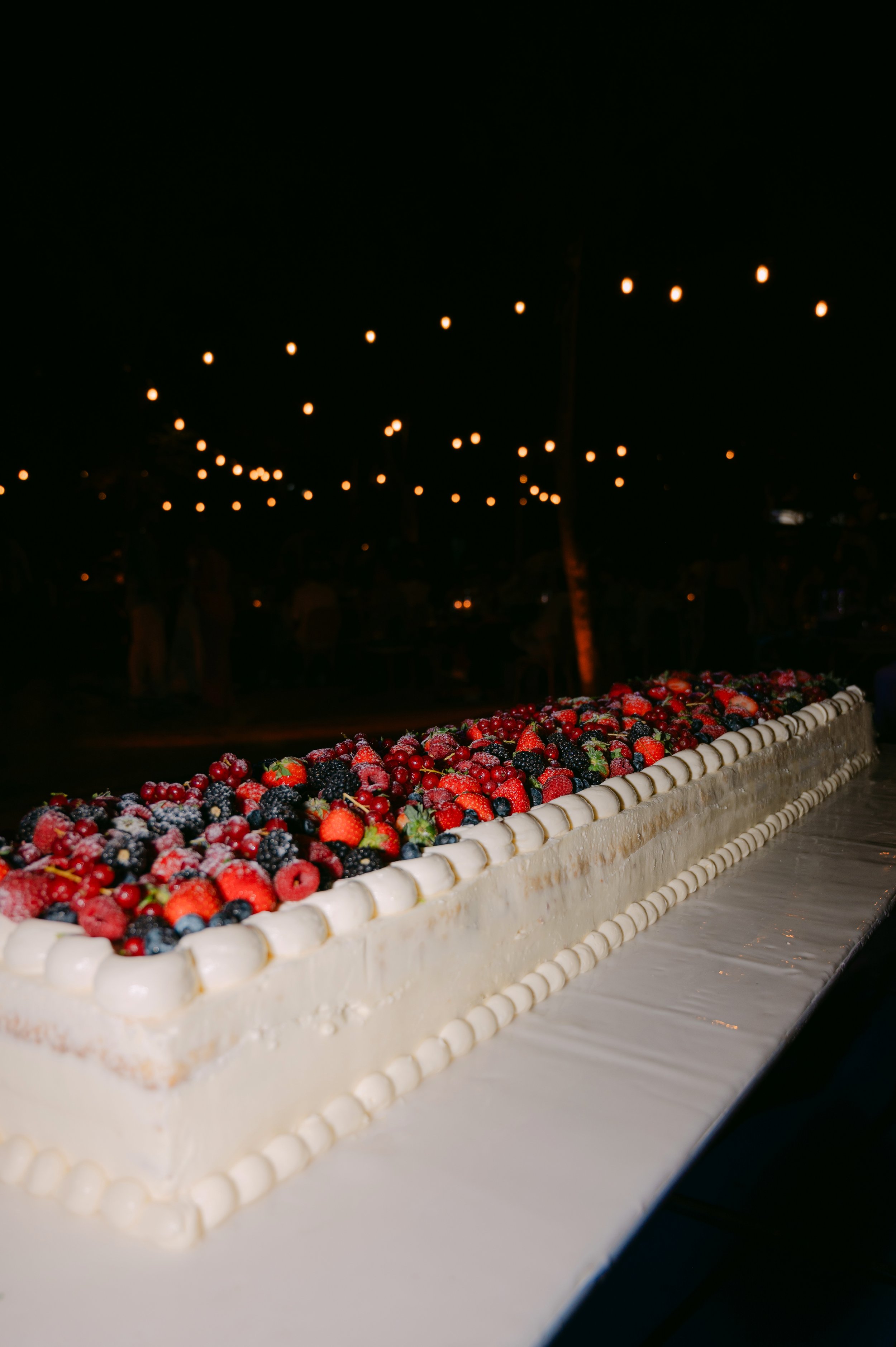 A long rectangular fruit-topped cake with white icing, decorated with mixed berries, on a table at night with string lights above.