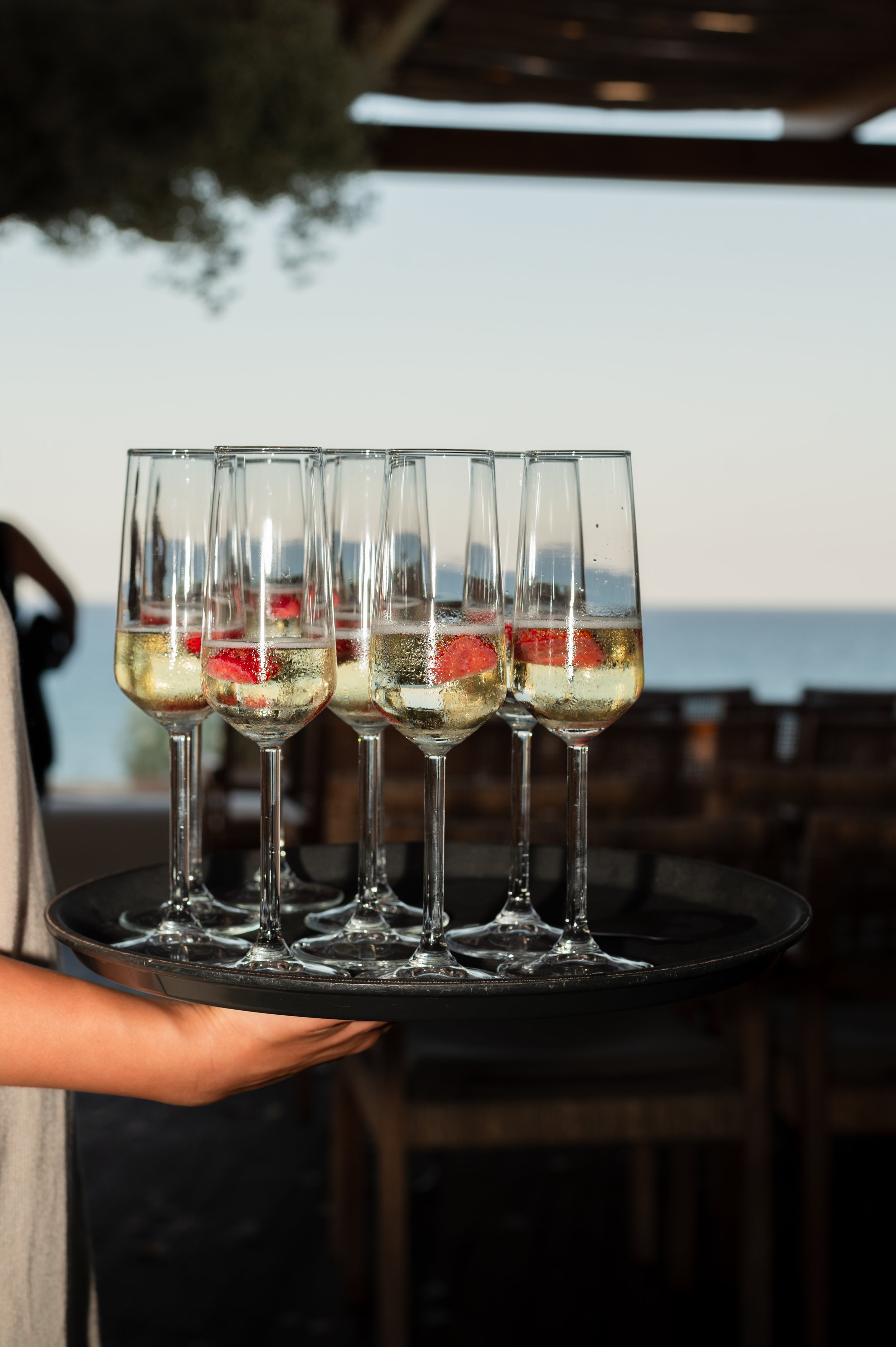 A person's hand holding a black tray with six glasses of sparkling wine garnished with strawberries, set outdoors with a view of the ocean in the background.