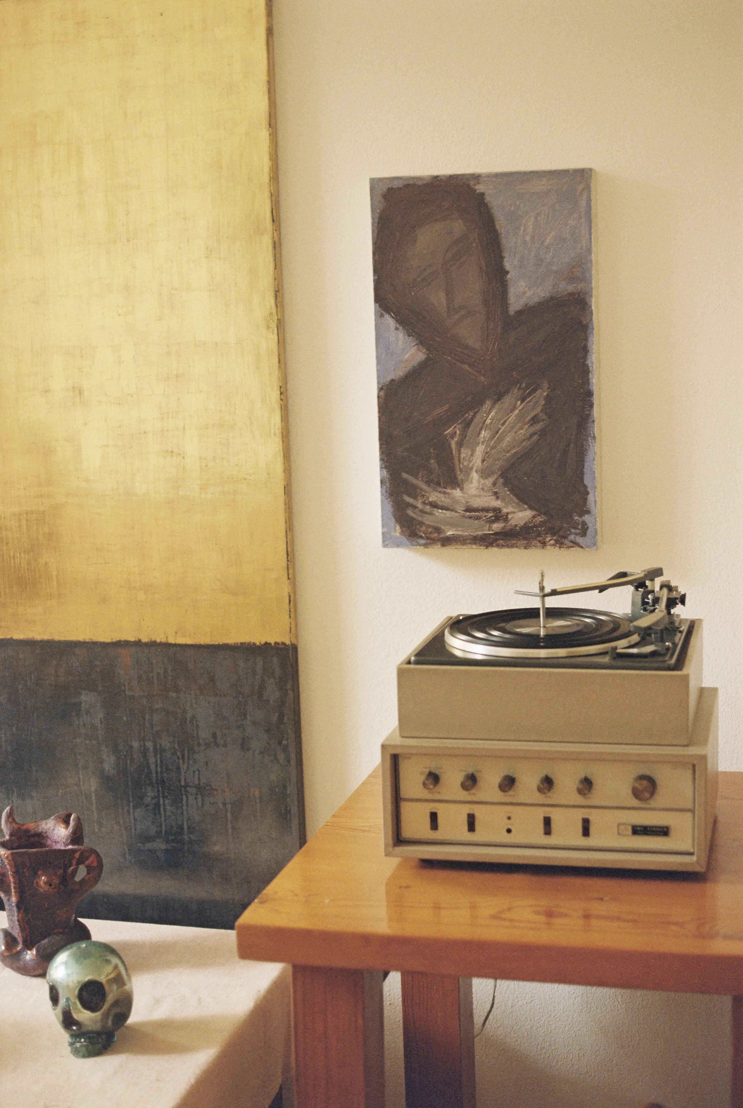 Vintage record player on a wooden table next to a skull-shaped object and a small figurine, with abstract art on the wall behind.