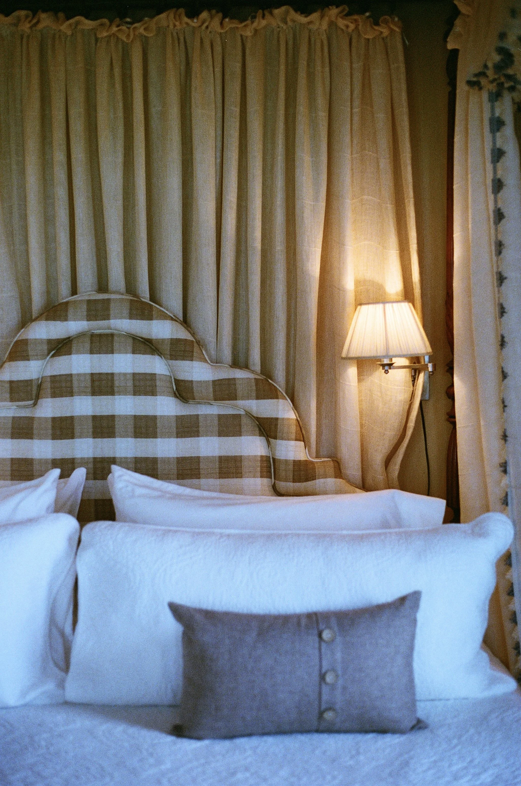 A bedroom with a bed featuring a plaid headboard, white blankets, and a gray pillow with buttons. Behind the bed are beige curtains, and to the right, there is a wall-mounted lamp with a pleated lampshade lighting up the area.