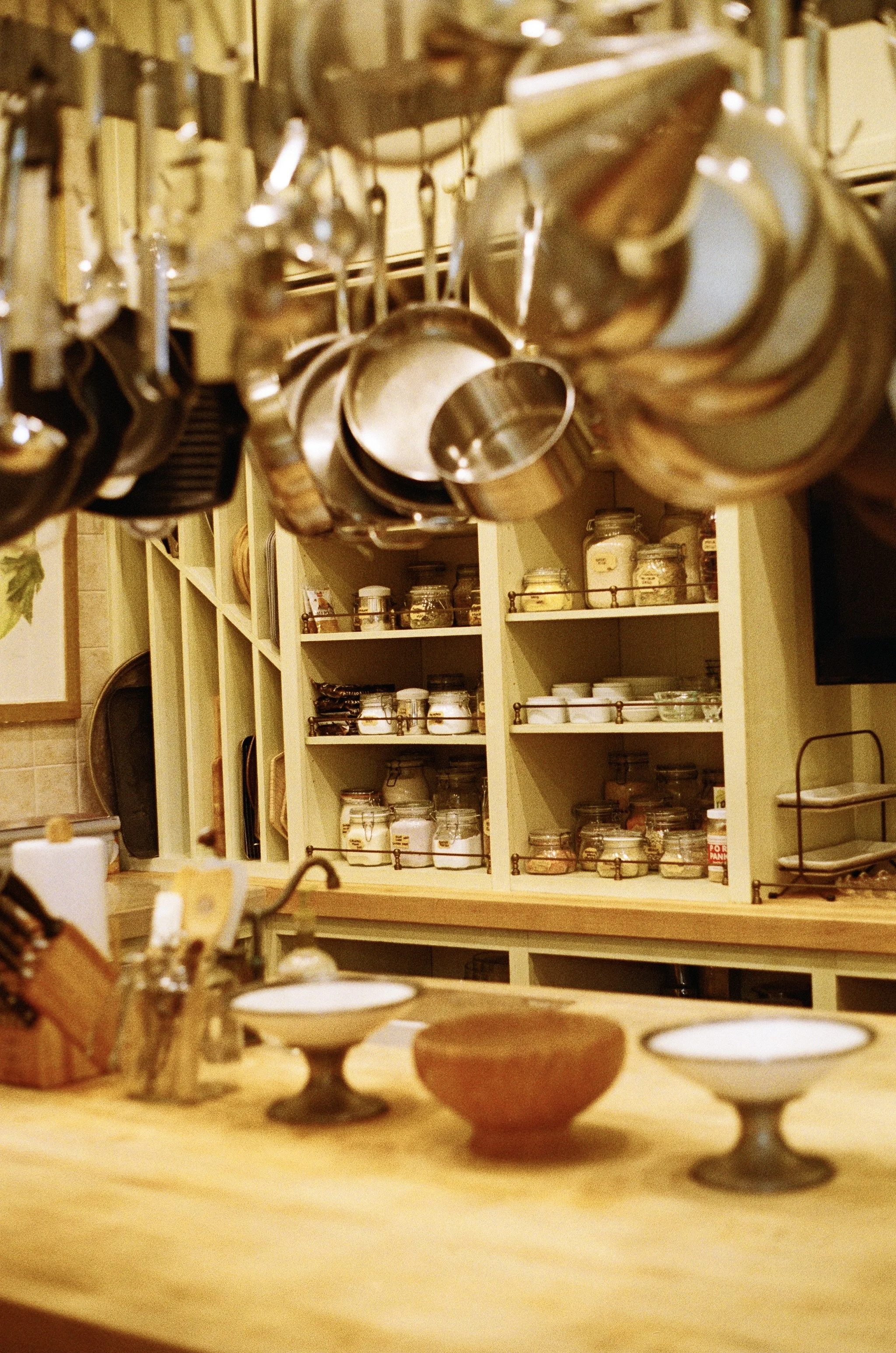 Kitchen with hanging metal pots and cups, shelves filled with jars and bowls, and a wooden counter with decorative bowls and utensils.