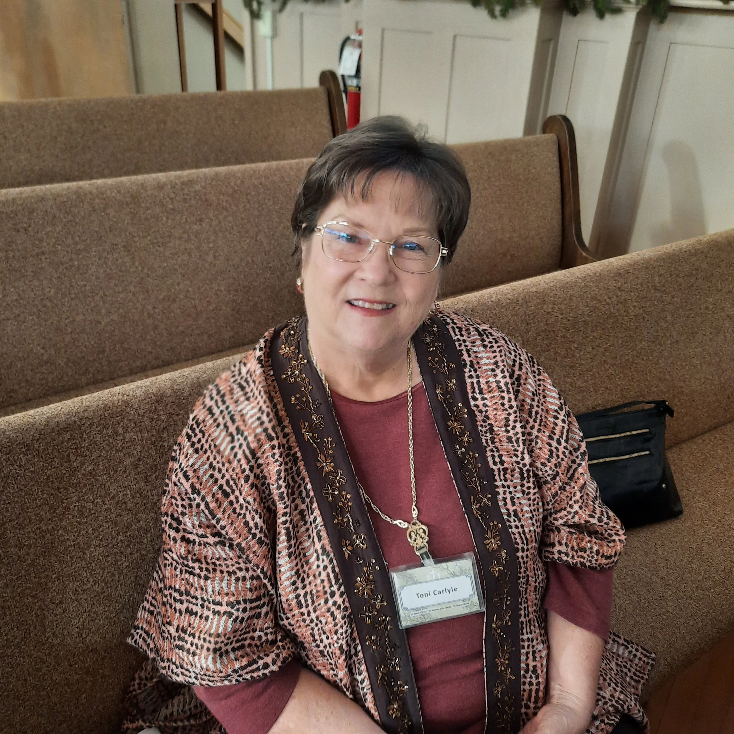 An older woman with short dark hair, glasses, and a warm smile sitting on a church pew. She is wearing a patterned shawl over a maroon top, with a name tag that reads "Toni Carlyle". Behind her, there are more church pews and a beige wall.