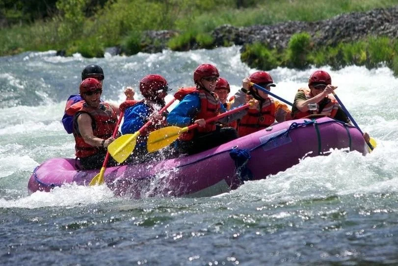 Six people in a pink inflatable raft wearing red helmets and life jackets, paddling through white water rapids on a river.