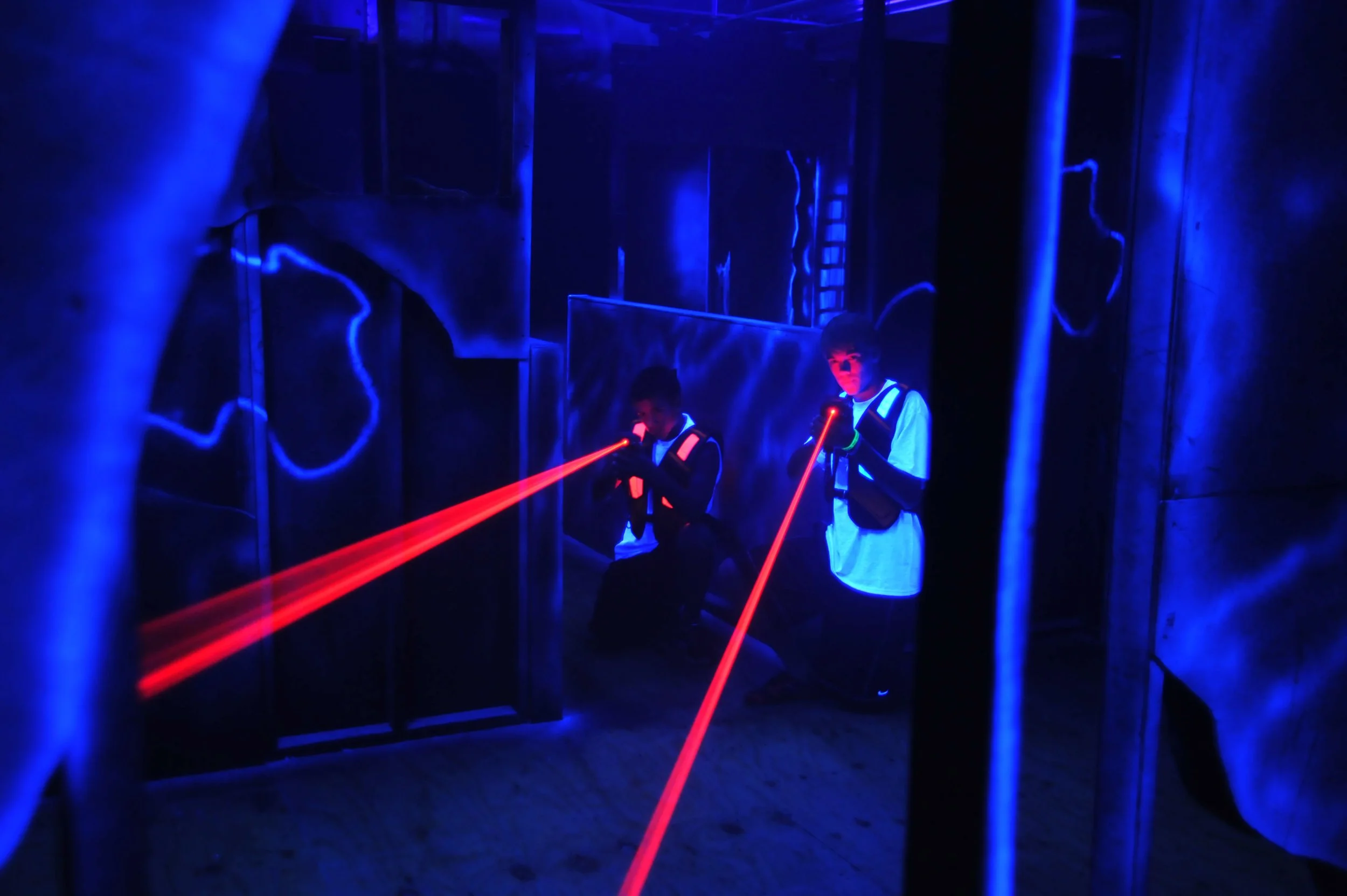 Two children in a dark room with blue lighting and mirror panels, playing with red laser pointers.
