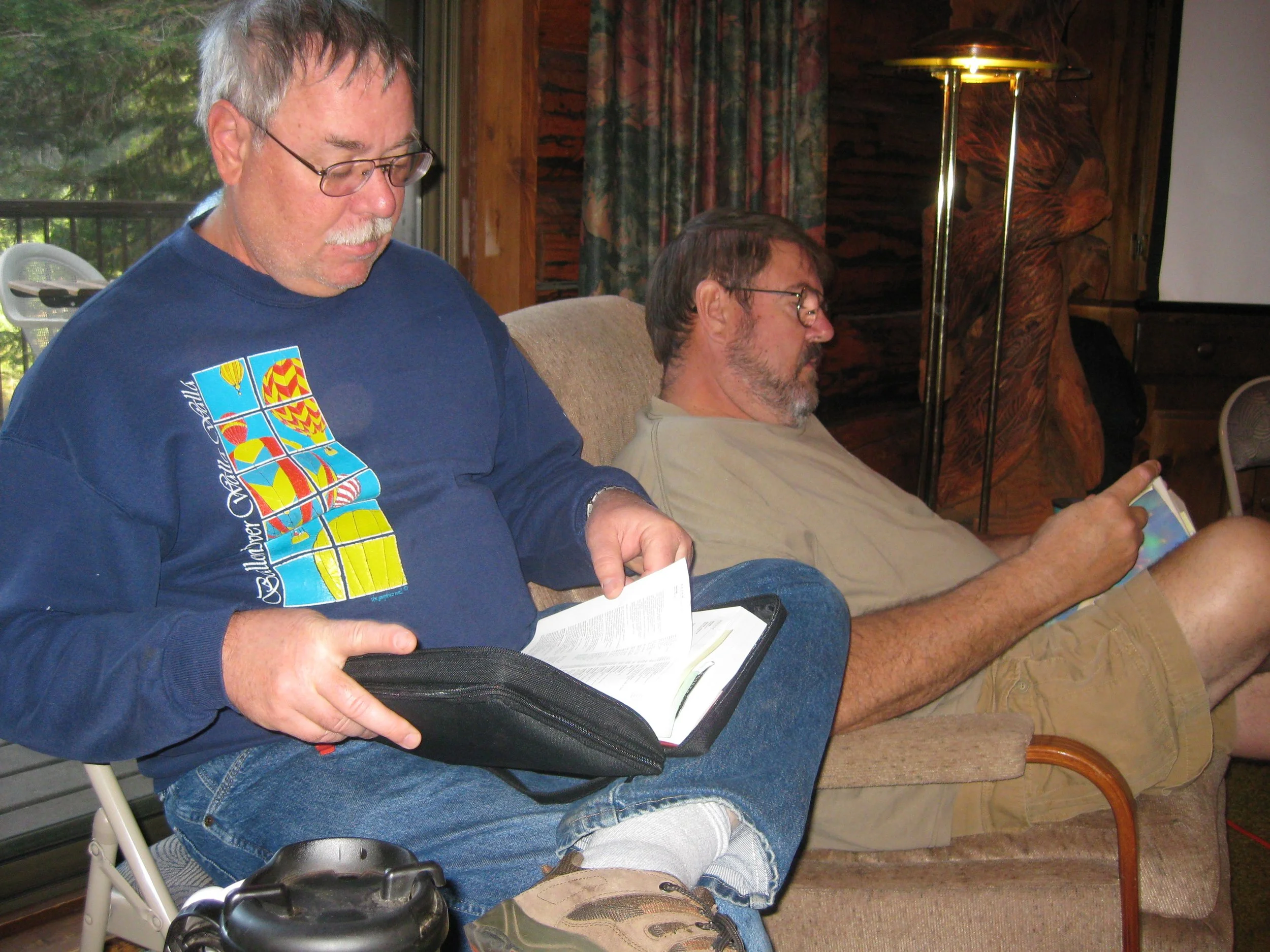 Two middle-aged men sitting on a couch indoors, both reading or looking at something in their hands; one is holding a book or folder, the other a tablet or phone.