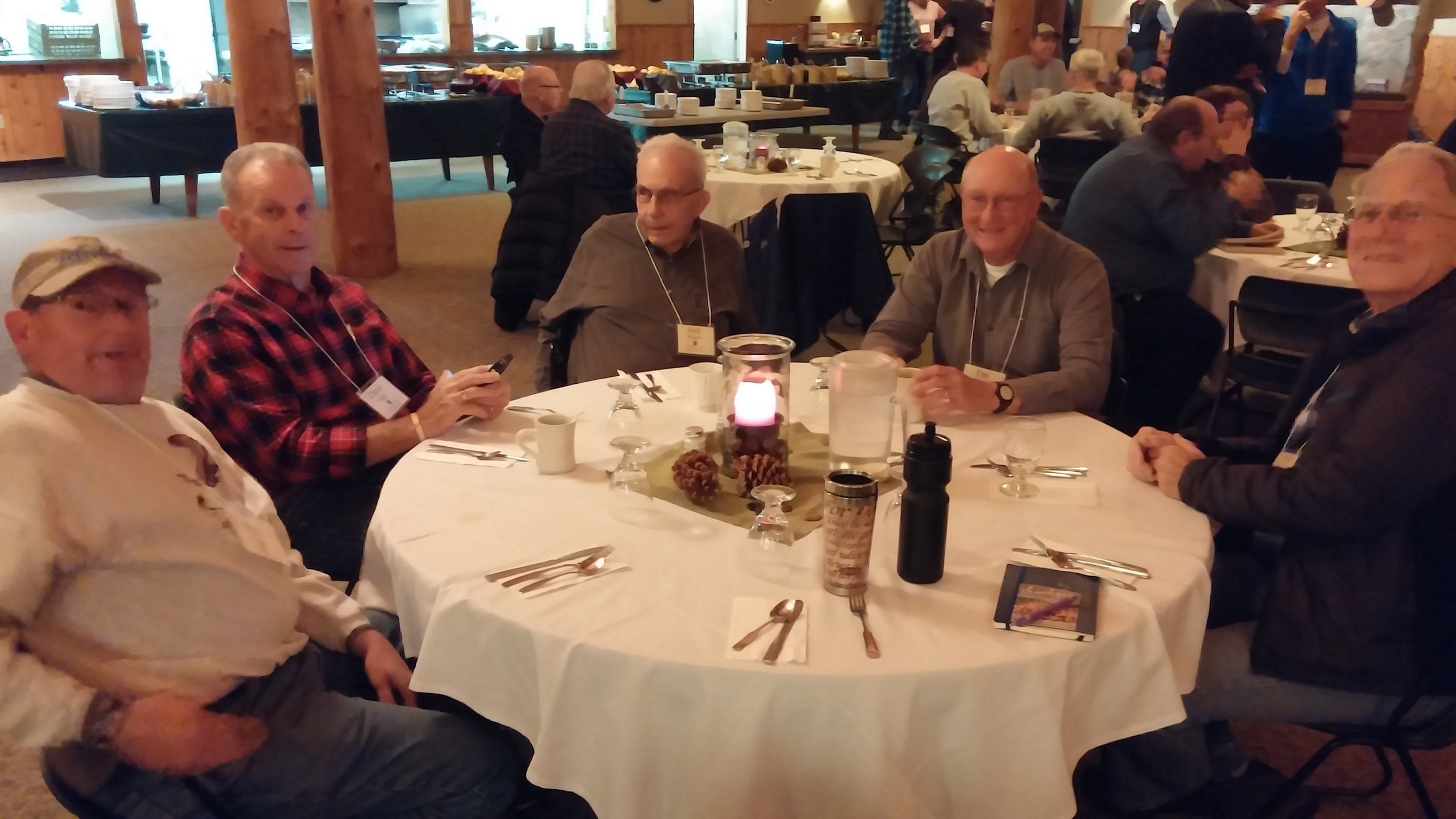 Five men sitting around a round dining table at a social event in a rustic, wood-paneled room.