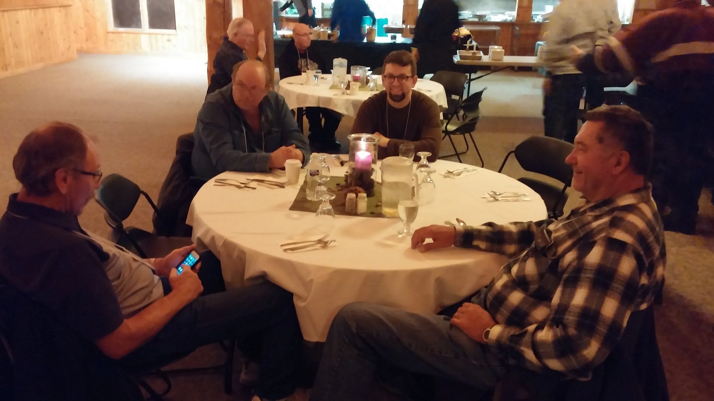 Four men sitting around a round table in a warmly lit dining area, one man looking at his phone, others smiling and chatting, with a candle and salt and pepper shakers on the table.
