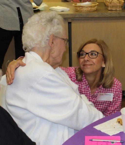 An elderly woman with white hair and glasses and a younger woman with blonde hair and glasses are sitting at a table, embracing and looking at each other warmly.