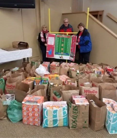 Three people stand behind a Christmas-themed Plinko game, with a large pile of grocery bags and boxes filled with donated food items in front of them, indicating a food drive or donation event.
