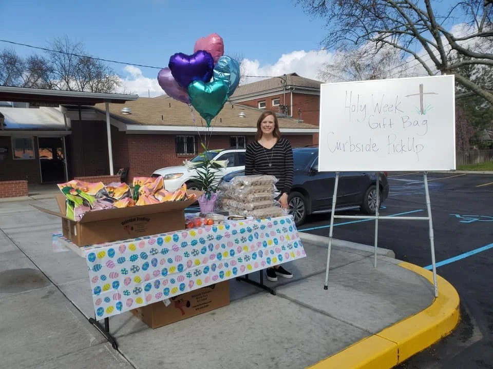 A girl selling gift bags and roses at an outdoor table decorated with an Easter-themed tablecloth. Balloons are tied to the table, and a whiteboard sign reads 'Holy Week Gift Bag, Myrtle Ficklep.'