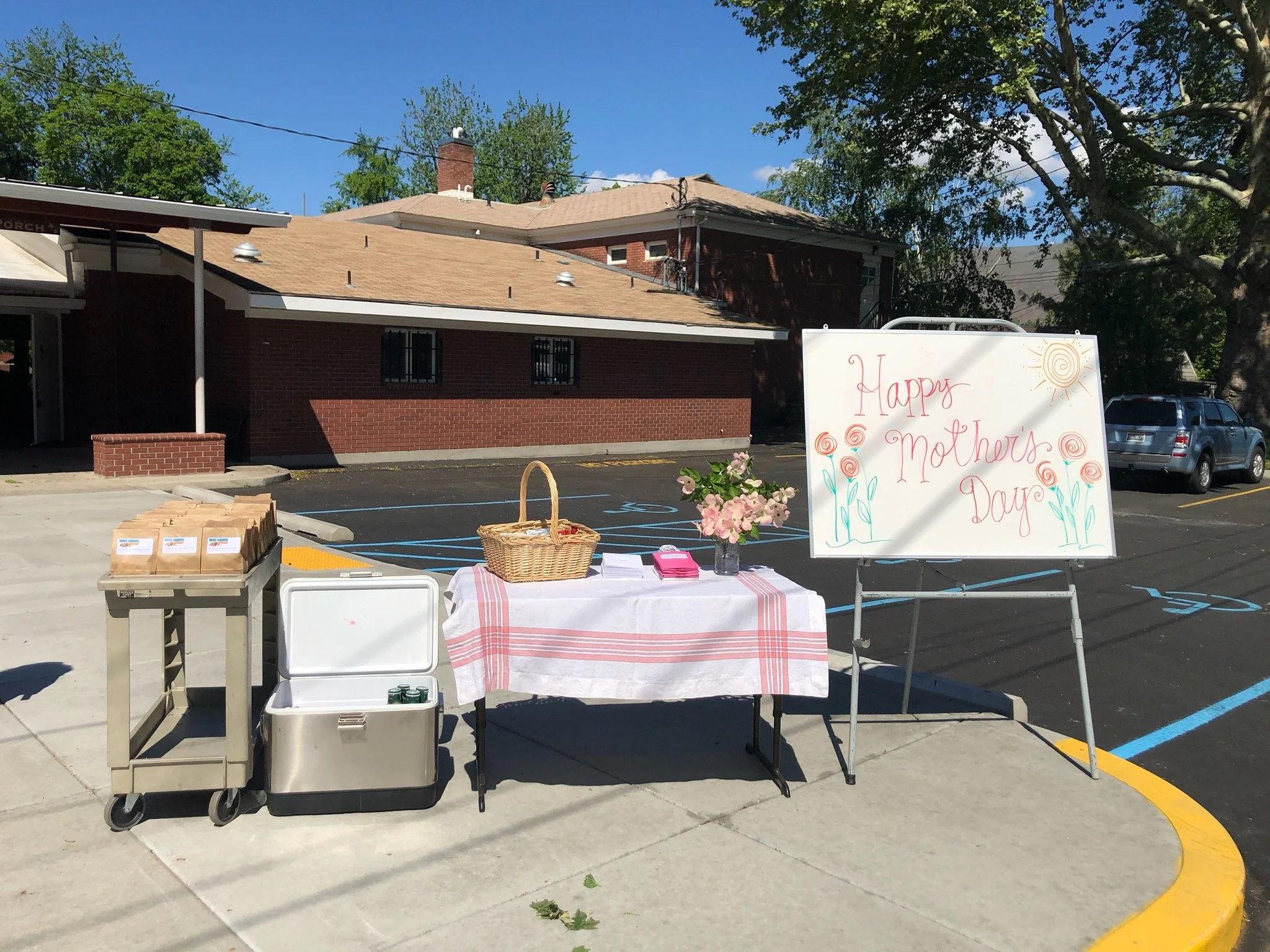 Table with flowers, a pink basket, and pink napkins next to a whiteboard with a "Happy Mother's Day" message, set up in a parking lot.