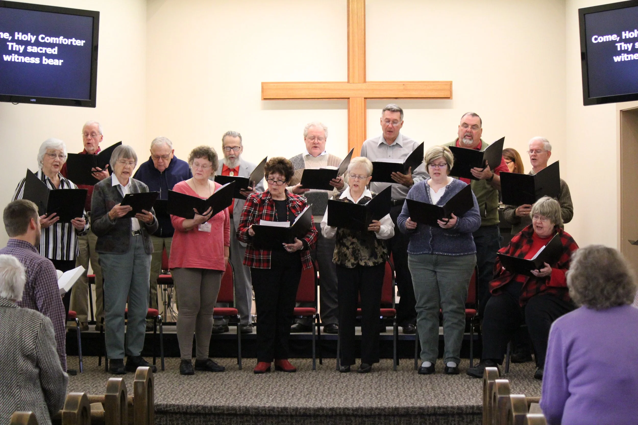 A choir of elderly men and women standing on risers in a church, singing from black folders. A large wooden cross hangs on the wall behind them. Several people are seated in the congregation, watching the performance.