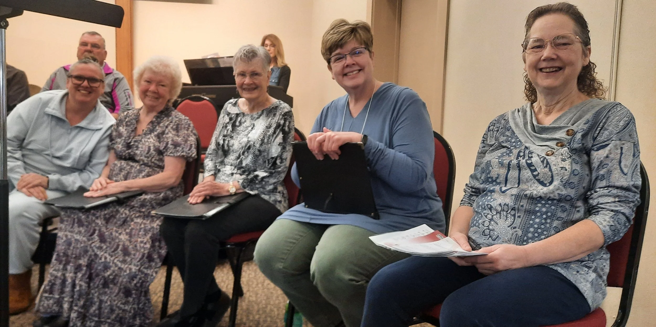 Group of five smiling women and two men sitting in a conference room, some holding notebooks, with a pianist and a woman at a music stand in the background.