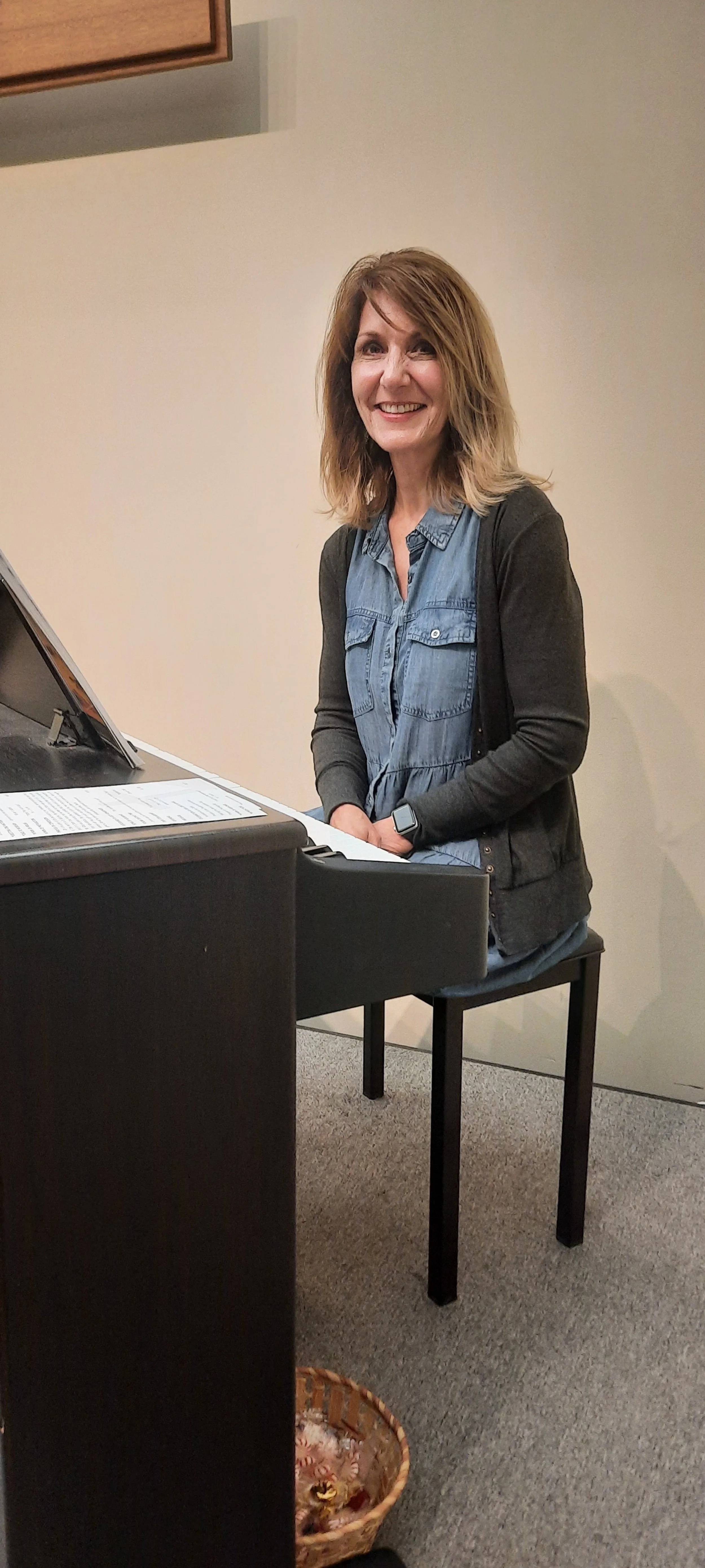 A woman with shoulder-length light brown hair, wearing a denim shirt and black cardigan, sitting and playing a black digital piano in a room with beige walls and gray carpet. She is smiling and looking at the camera.