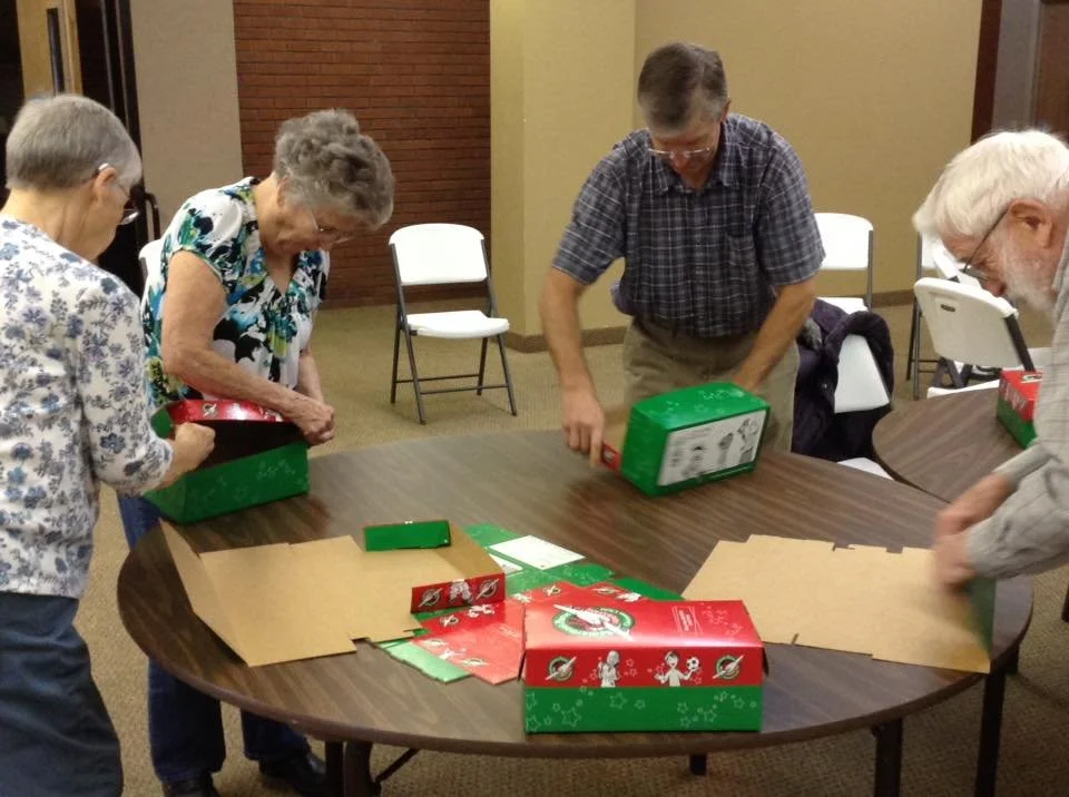 Four elderly people standing around a table unboxing Christmas-themed gift boxes. There are empty chairs in the background.
