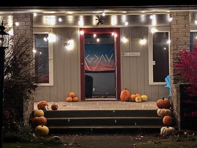Front porch decorated with pumpkins, surrounded by string lights, leading to a house with gray siding and a partially covered sitting area.