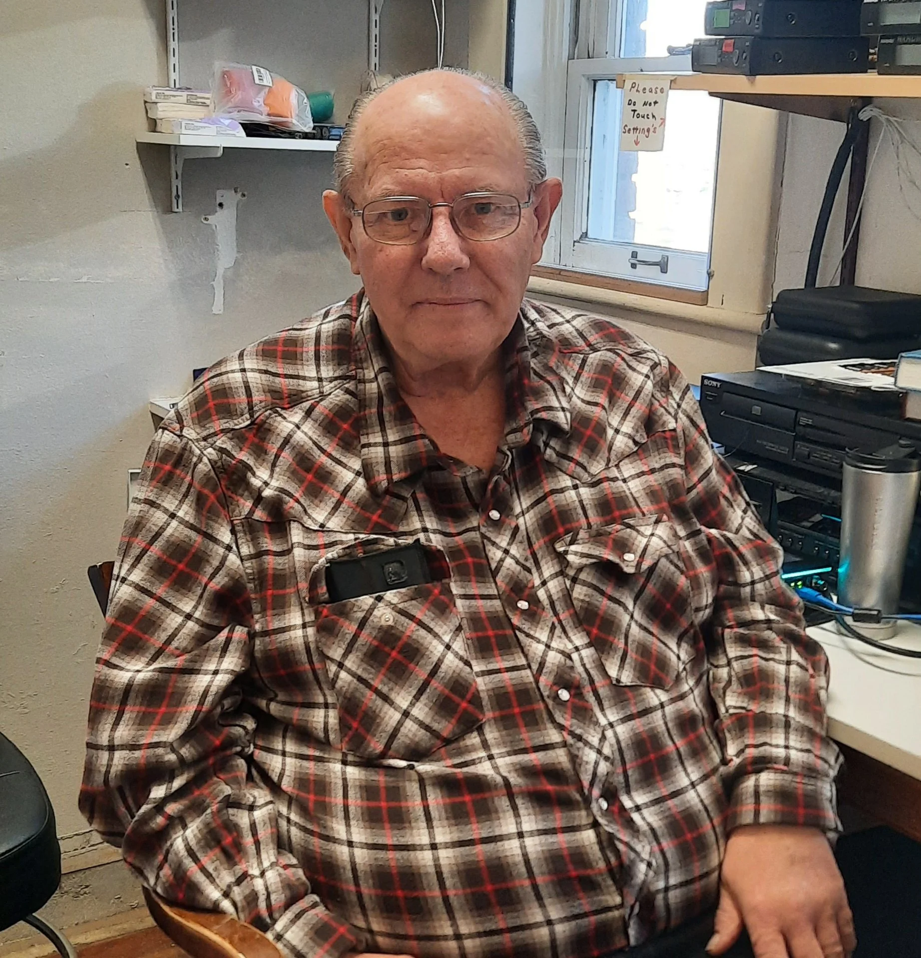 An elderly man wearing glasses and a red, black, and white plaid shirt sitting at a desk in an office or workshop. There are shelves with supplies and equipment, a window with a sign saying 'Please Do Not Touch Settings,' and electronic devices on th