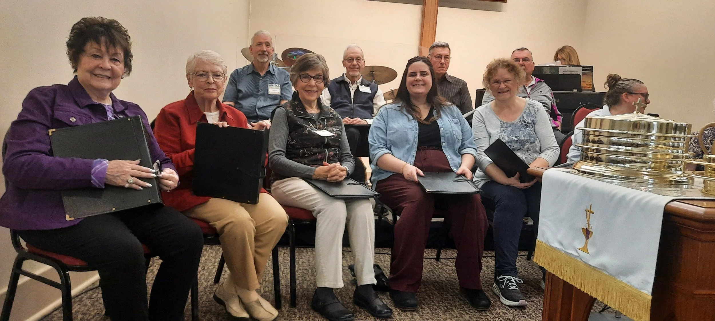 Group of nine people seated and standing in a room, holding hymnals or folders, with a table featuring seated golden communion trays on the right.